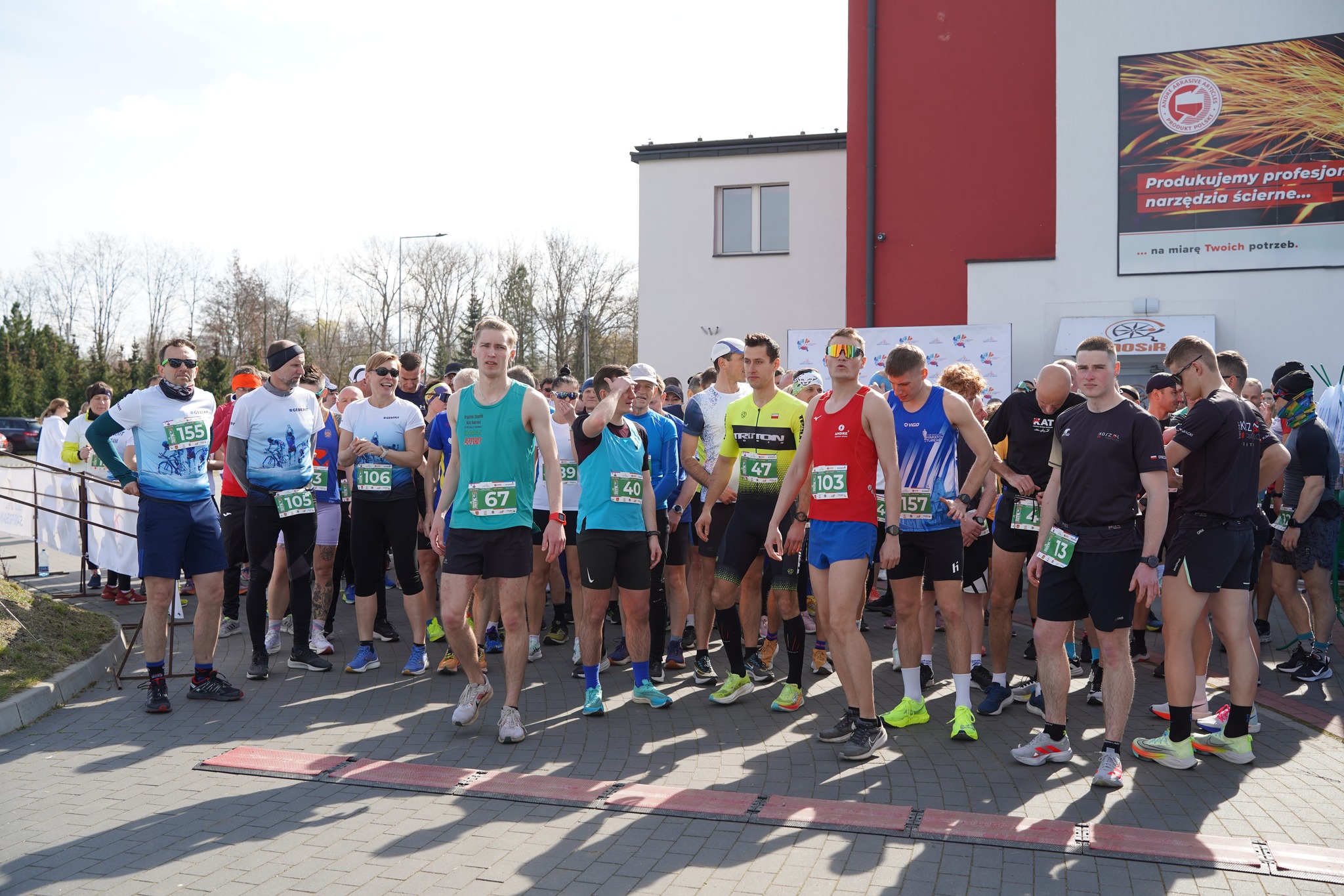 Zdjęcie przedstawia grupę biegaczy na linii startu zawodów ulicznych. Uczestnicy są w sportowych strojach, z numerami startowymi, gotowi do rozpoczęcia biegu. w tle widać budynek z banerem oraz logo MOSiR. Pogoda jest słoneczna.
