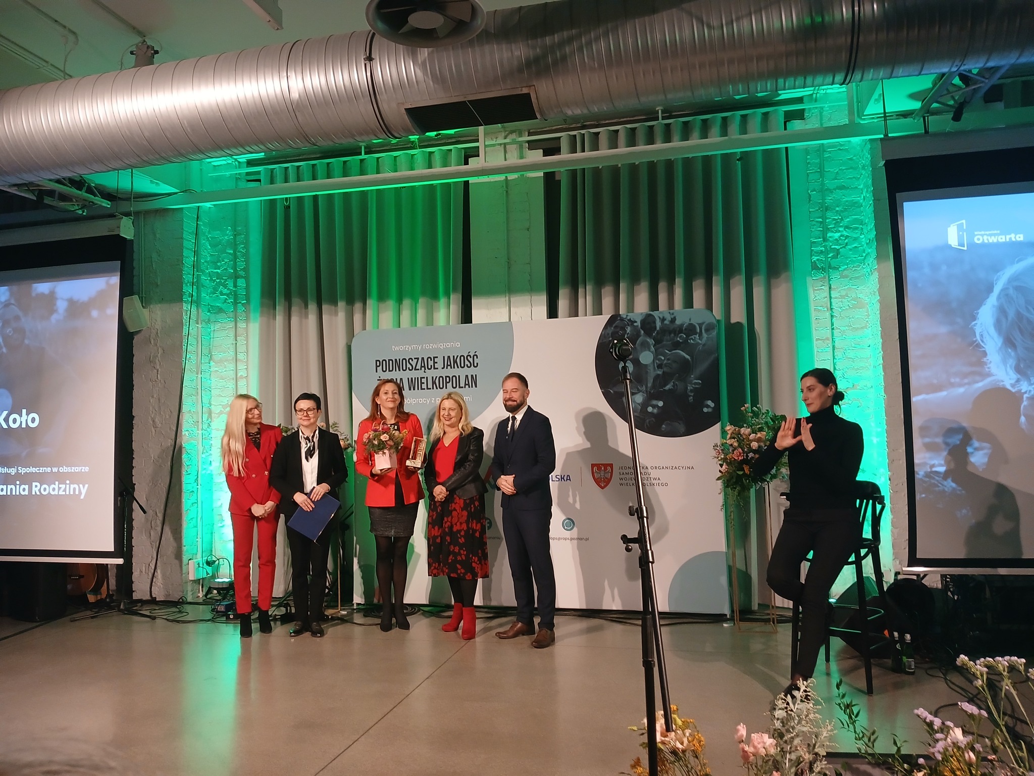 Zdjęcie z gali wręczania nagród. Na środku znajduje się grupa osób, w tym kobieta z bukietem kwiatów i statuetką, laureatka wyróżnienia. Po jej bokach stoją inne osoby, najprawdopodobniej współorganizatorzy lub współlaureaci.