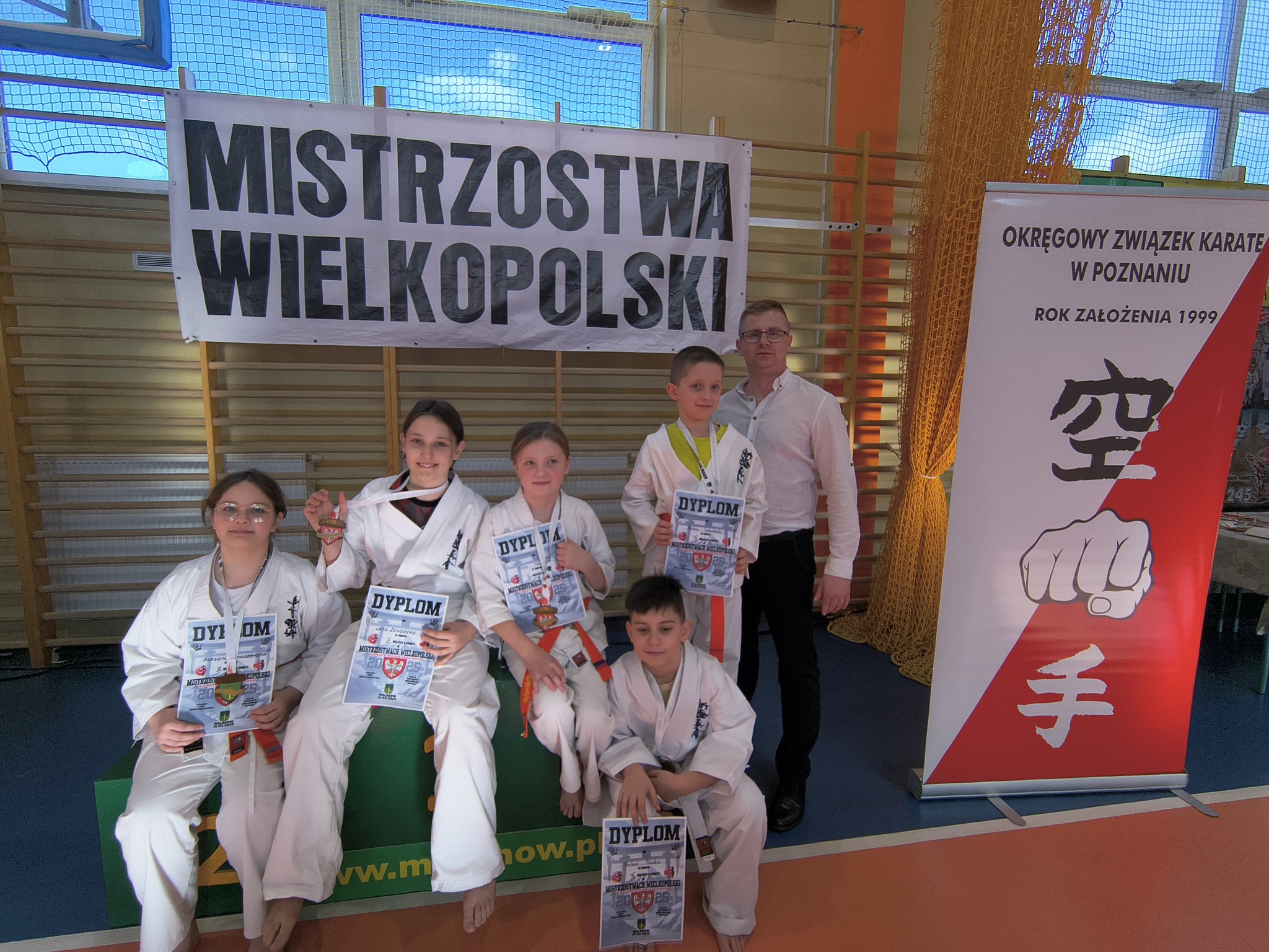 Grupa młodych karateków, dziewcząt i chłopców z dyplomami i medalami, pozuje do zdjęcia w hali sportowej z opiekunem. Za nimi widać drabinki do ćwiczeń, na nich wiszący baner a z prawej strony roll-up Okręgowego Związku Karate
