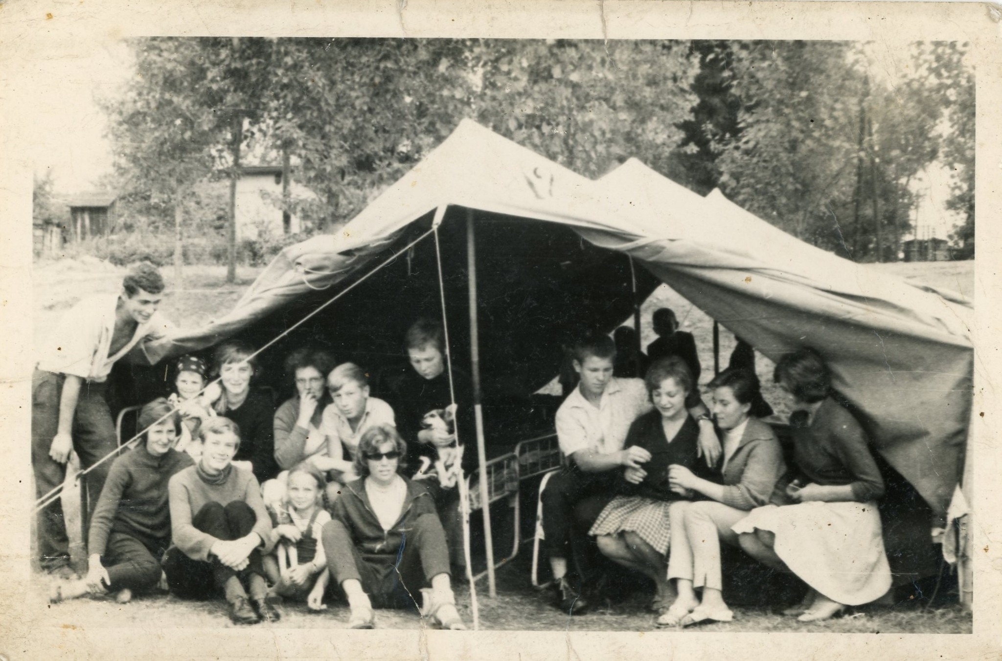 Na zdjęciu widać grupę młodych osób siedzących i klęczących przed dużym namiotem w plenerze. To letni obóz — wszyscy są ubrani swobodnie, a wokół widać drzewa i trawiasty teren. Zdjęcie jest czarno-białe.