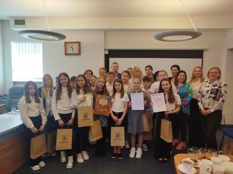 Grupowe zdjęcie laureatów konkursu recytatorskiego zrobione w czytelni Miejskiej i Powiatowej Biblioteki Publicznej w Kole. Dziewczęta w pierwszym rzędzie trzymają w dłoniach dyplomy i torebki z upominkami z herbem miasta Koła.