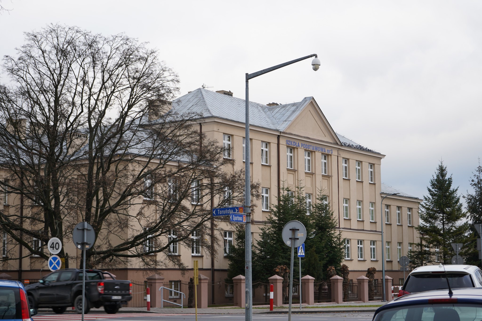 Na zdjęciu widoczny jest budynek szkoły oznaczonej jako Szkoła Podstawowa nr 3. Jest to duży, jasnobeżowy budynek z wieloma oknami i charakterystycznym dachem pokrytym blachą. Przed budynkiem znajdują się drzewa bez liści, co sugeruje porę jesienną.