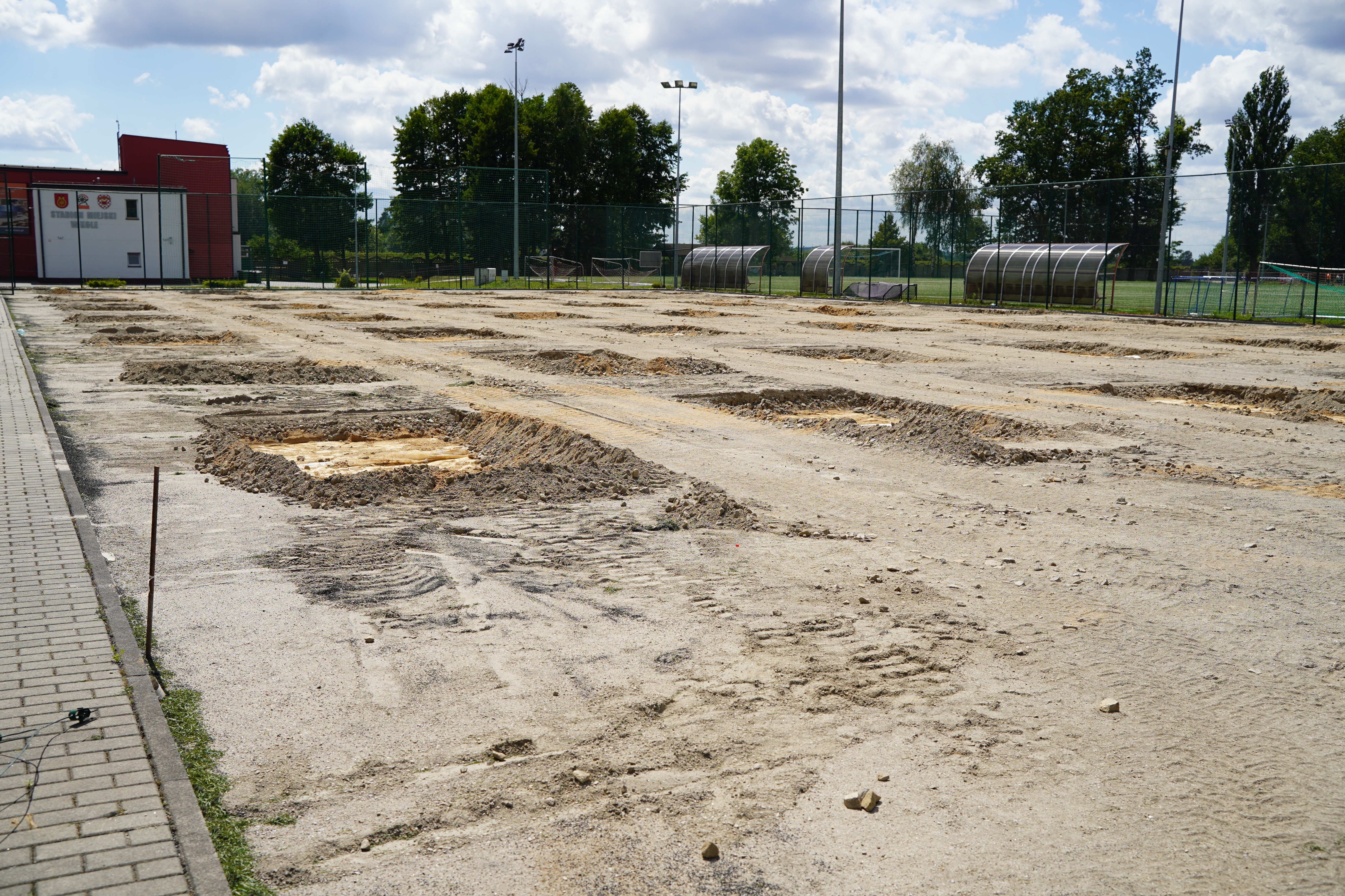 Na zdjęciu widać teren w trakcie remontu – usunięto starą nawierzchnię, a na placu znajdują się wykopy. W tle widać boiska sportowe i budynek stadionu.