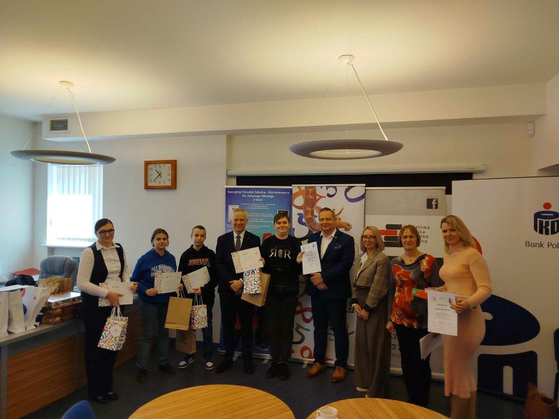 Grupowe zdjęcie laureatów Miejskiej Olimpiady Ekonomicznej SKO zrobione w czytelni Powiatowej i Miejskiej Bibliotece Publicznej w Kole. Laureaci trzymają w dłoniach nagrody. Wśród nich znajdują się osoby dorosłe.