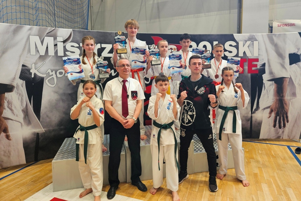 Grupa młodych zawodników karate pozujących z medalami i dyplomami po zawodach, wraz z trenerem i sędzią, na tle baneru turnieju.