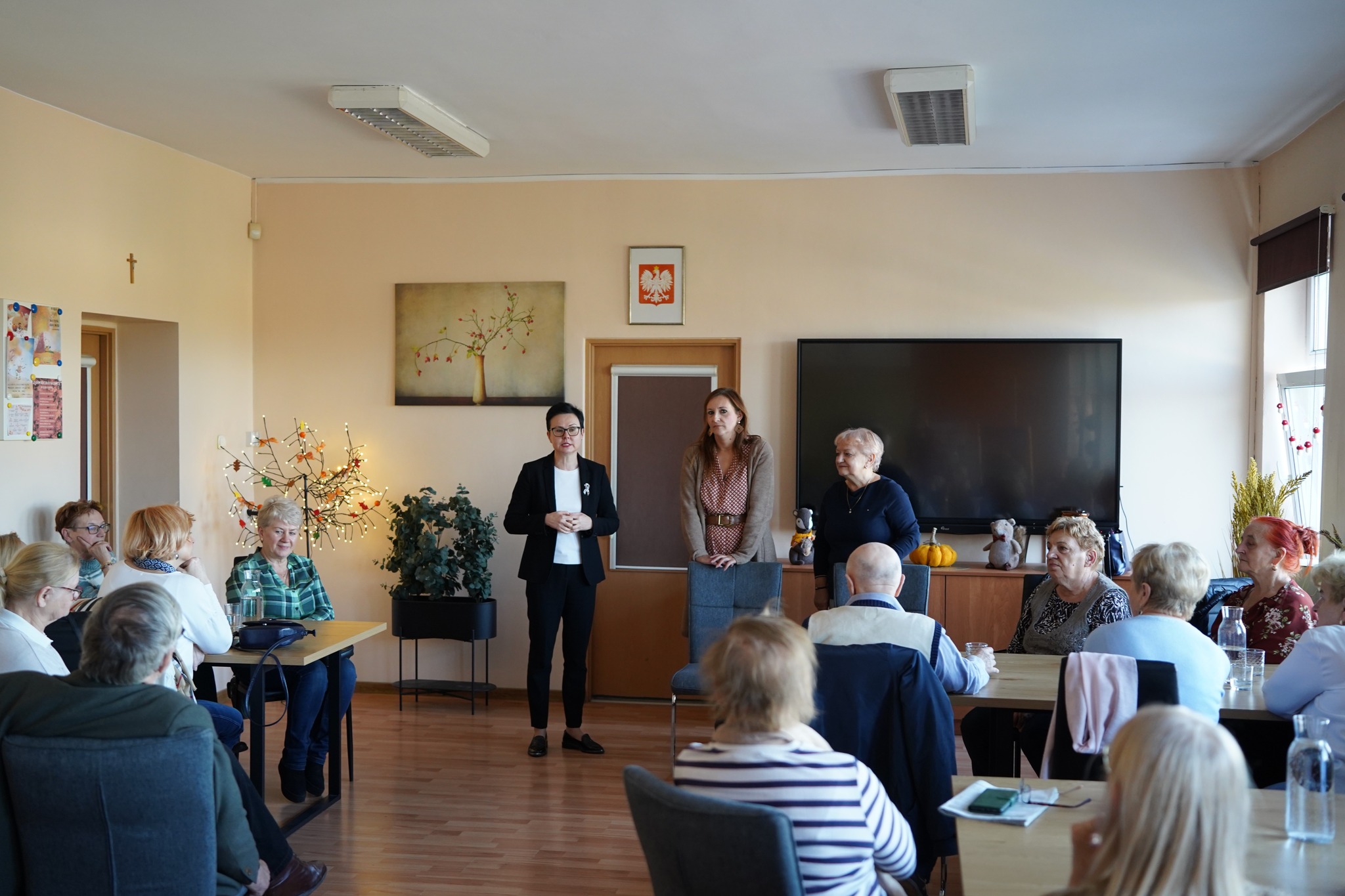 Zdjęcie przedstawia spotkanie grupy osób w sali — kilka osób stoi i przemawia, a reszta siedzi przy stołach, słuchając.