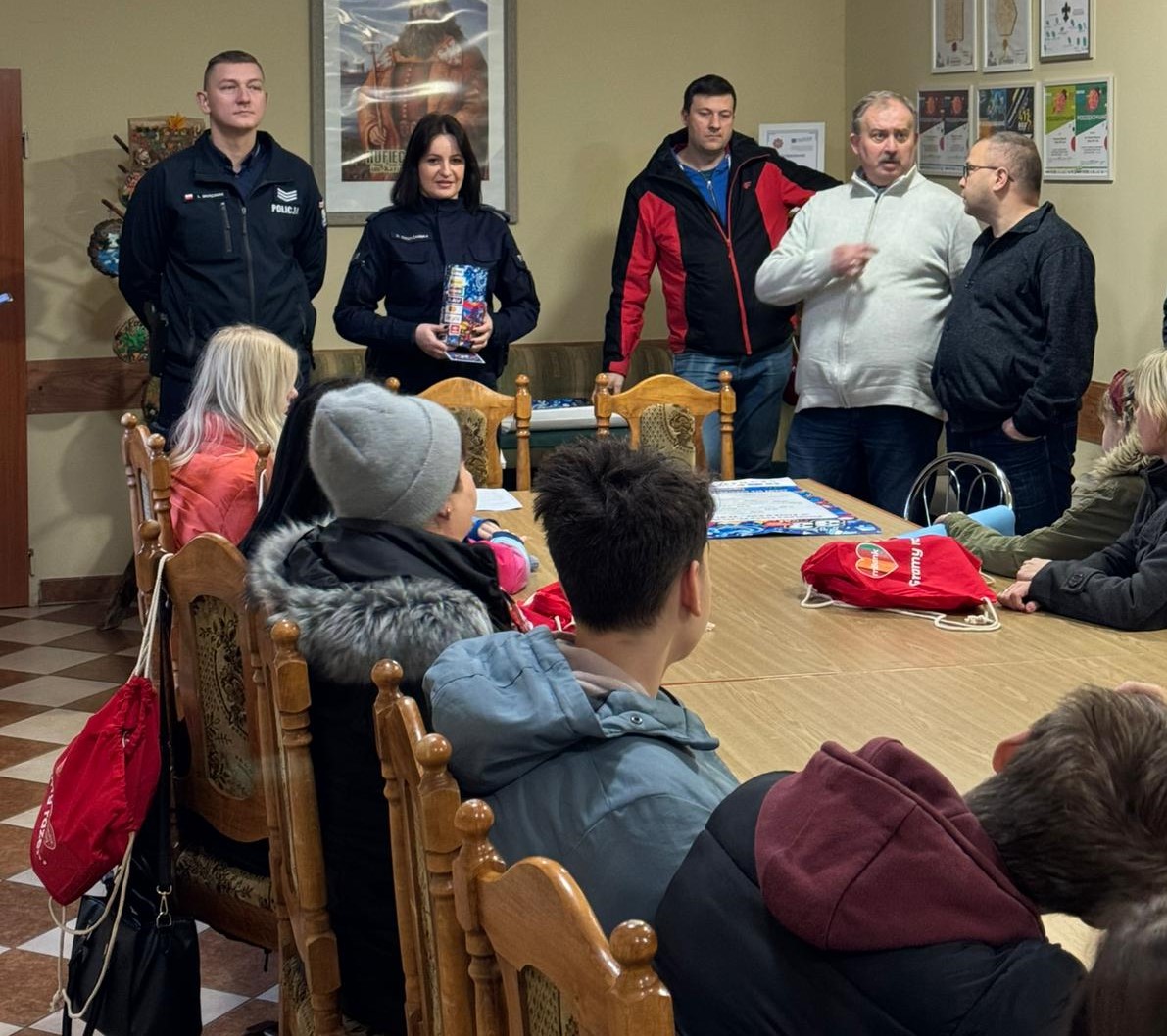 Na zdjęciu widać grupę ludzi w sali, siedzących przy stole oraz kilku stojących, w tym dwie osoby w mundurach policyjnych.