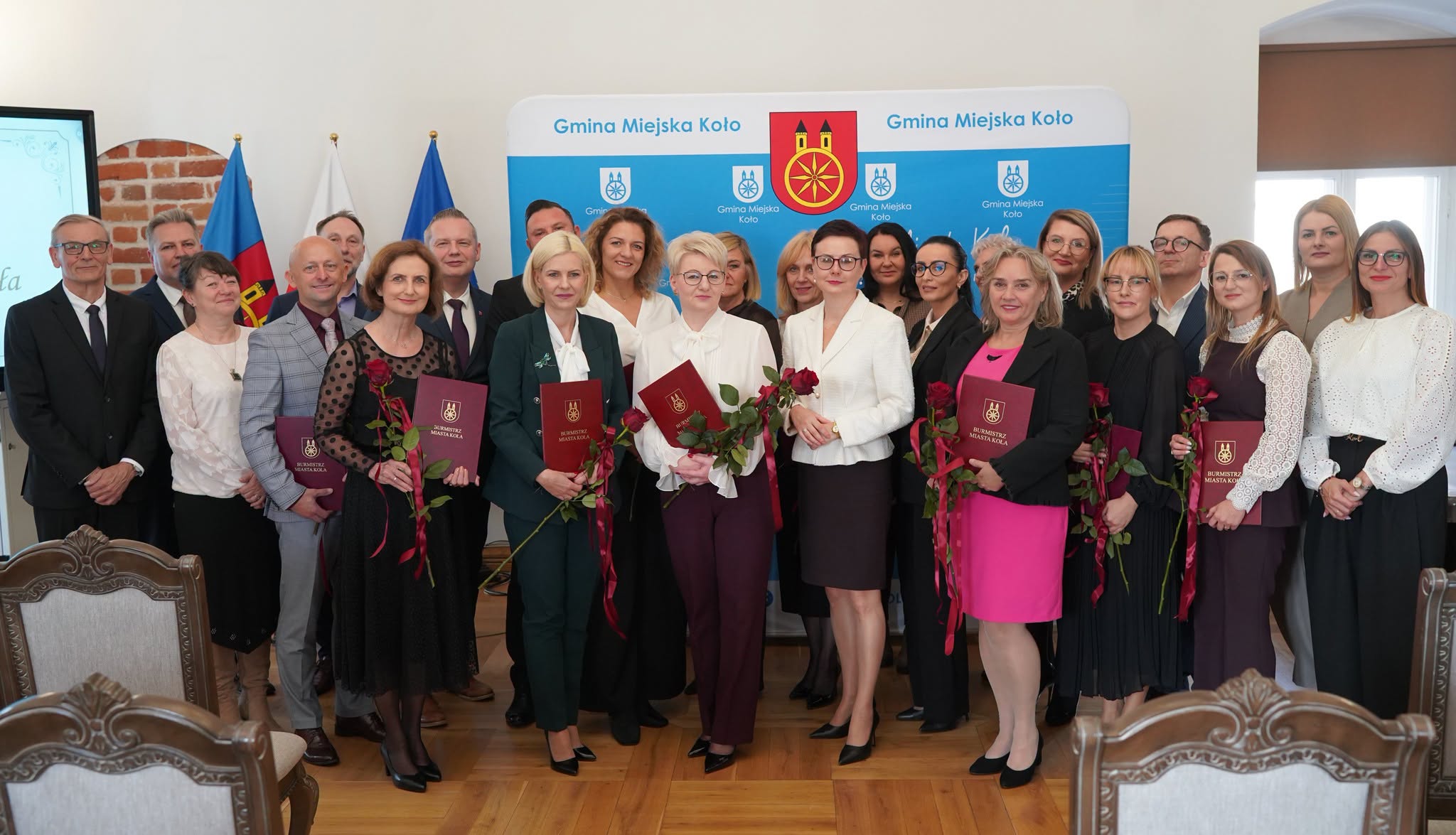 Grupa około 30 osób, głównie kobiet w eleganckich strojach, pozuje w sali z drewnianą podłogą na tle niebieskiej ścianki z herbem i napisem Gmina Miejska Koło. Część trzyma bordowe teczki z napisem Burmistrz Miasta Koła i czerwone róże. W tle flagi.