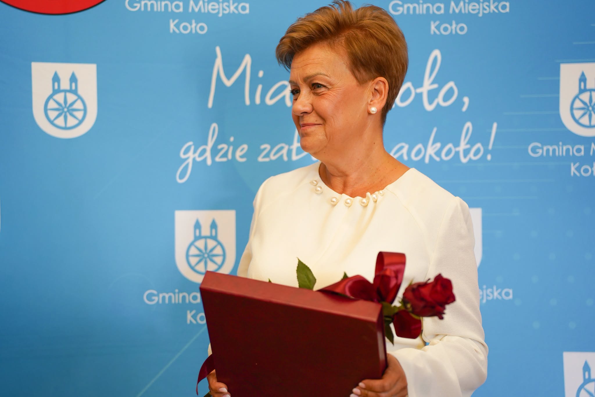 Uśmiechnięta kobieta o krótkich włosach, ubrana w kremową sukienkę z perełkami przy dekolcie, trzyma bordową teczkę i bukiet czerwonych róż z wstążką. Patrzy w bok. W tle niebieski baner z herbem Koła i hasłem Miasto Koło, gdzie zabytki wokoło!.