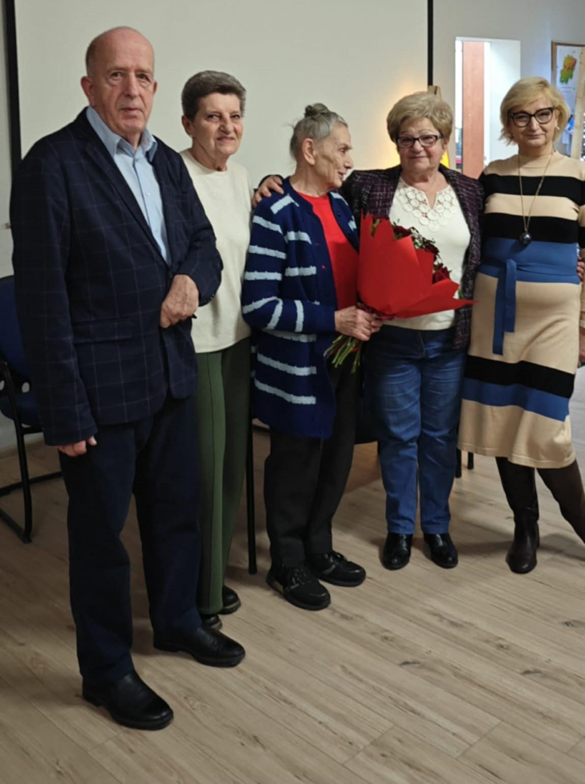 Na zdjęciu widać grupę sześciu starszych osób stojących obok siebie w sali. Jedna z kobiet trzyma bukiet czerwonych kwiatów, a cała grupa wygląda na uczestników uroczystości lub spotkania okolicznościowego.