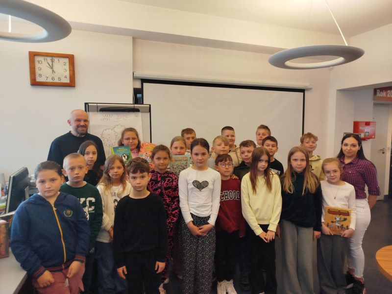 Grupowe zdjęcie uczestników spotkania z Tomaszem Samojlikiem, autorem komiksów i książek dla dzieci, zrobione w czytelni Powiatowej i Miejskiej Bibliotece Publicznej w Kole. W tle widać ekran wiszący na ścianie.