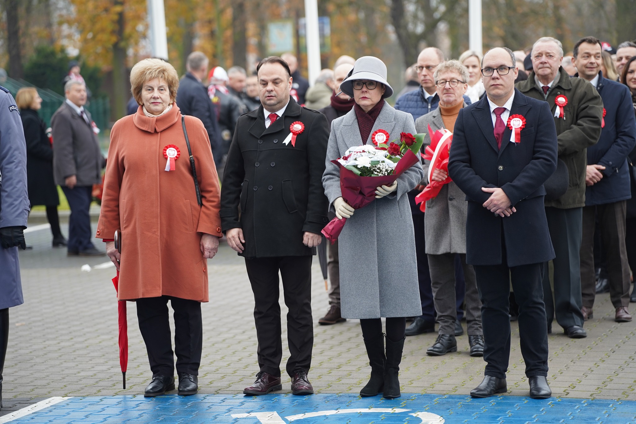 Na zdjęciu widać grupę osób uczestniczących w uroczystości patriotycznej. W pierwszym rzędzie stoją cztery osoby z biało-czerwonymi kokardami, a jedna z nich trzyma bukiet kwiatów. W tle widoczny jest tłum uczestników oraz jesienne drzewa.