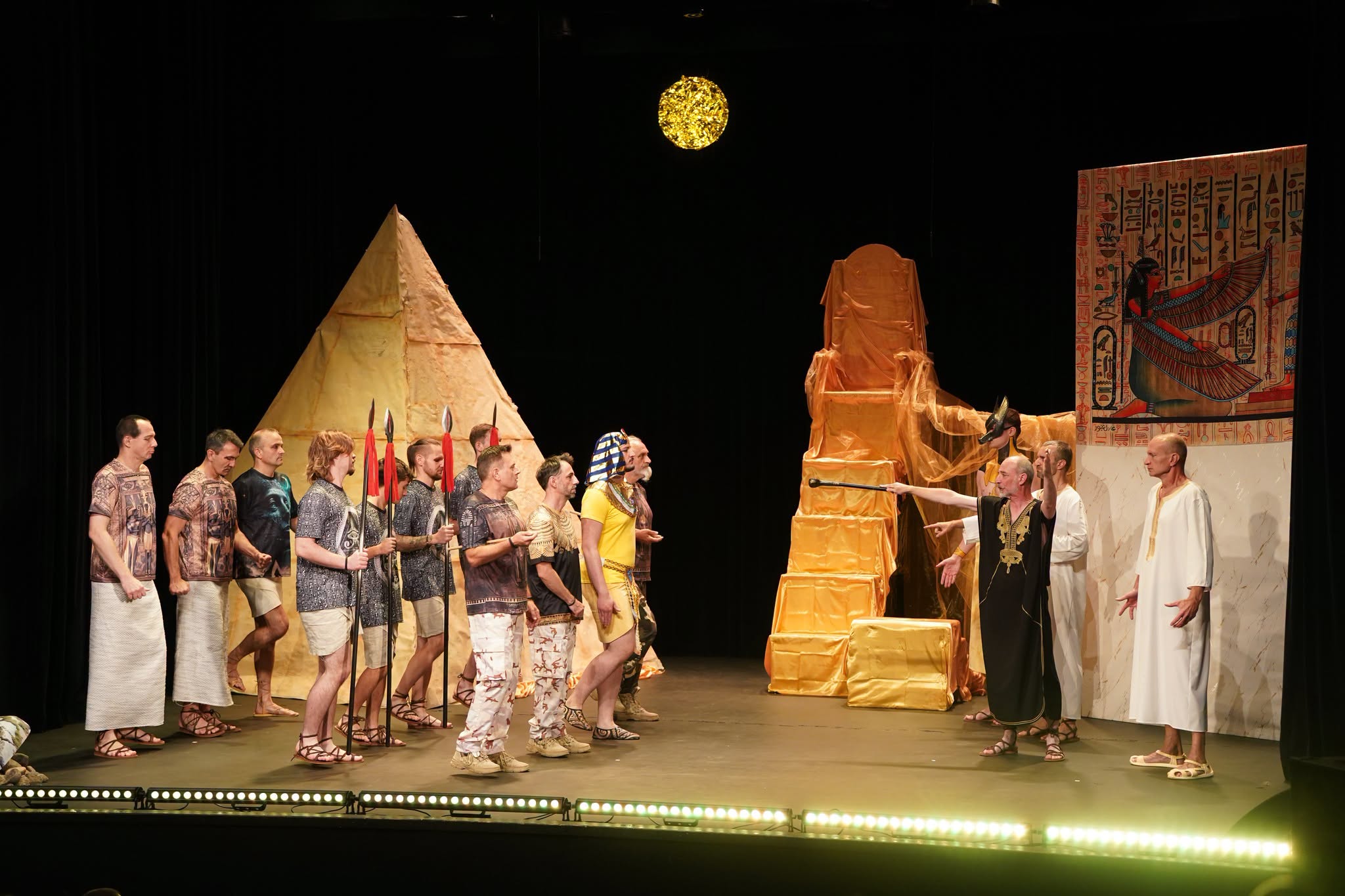 Scena teatralna: grupa aktorów w strojach starożytnych Egipcjan, część z włóczniami, stoi po lewej. Po prawej mężczyzna w czarnej tunice wskazuje laską na postać w białej szacie. W tle tekturowa piramida, złoty tron, papirus z boginią i złote słońce.