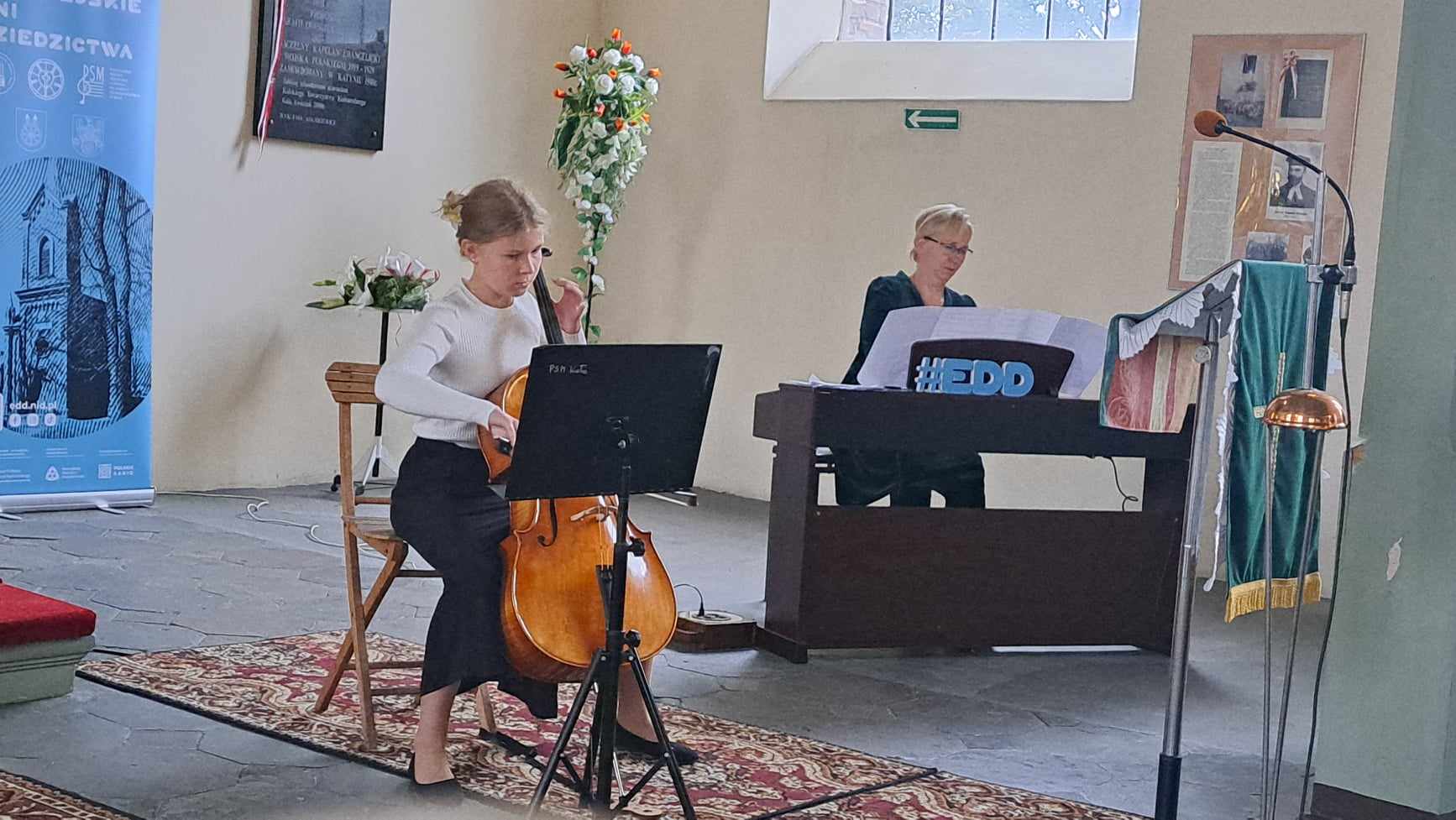 Zdjęcie przedstawia koncert w kościele ewangelickim w Kole. Kobieta gra na wiolonczeli, a w tle inna osoba gra na klawiszach. Na ścianie wisi plakat z napisem Dziedzictwa oraz kwiaty obok.