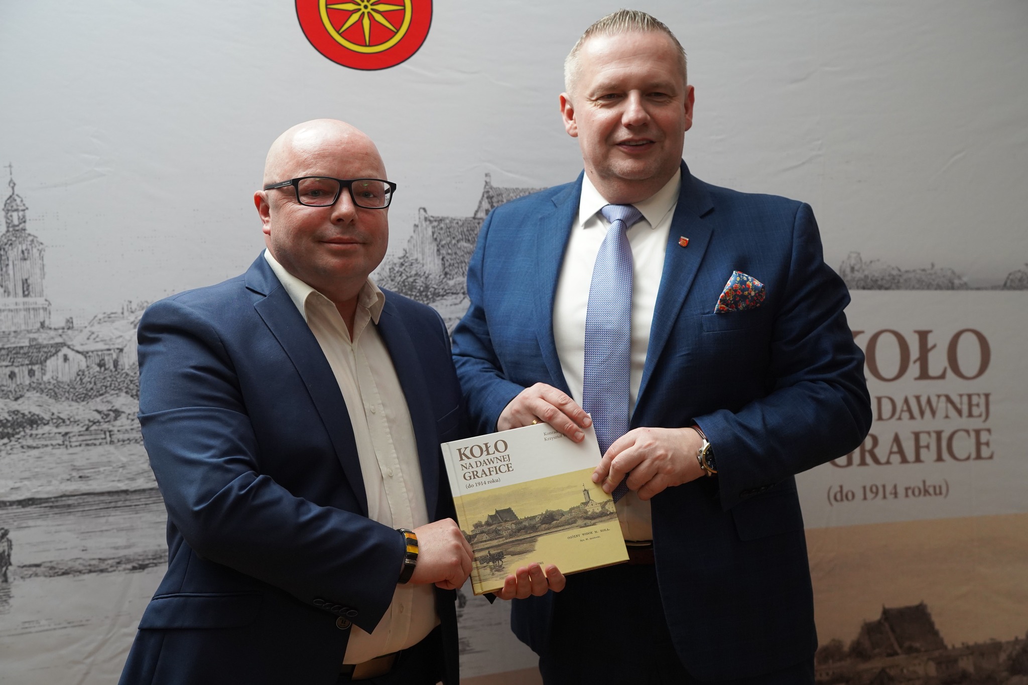 Na zdjęciu widać dwóch mężczyzn w garniturach, którzy stoją obok siebie i pozują do fotografii. Obaj trzymają wspólnie w dłoniach egzemplarz publikacji, z wyraźną dumą i zadowoleniem. W tle znajduje się ścianka promocyjna z grafiką i napisami.