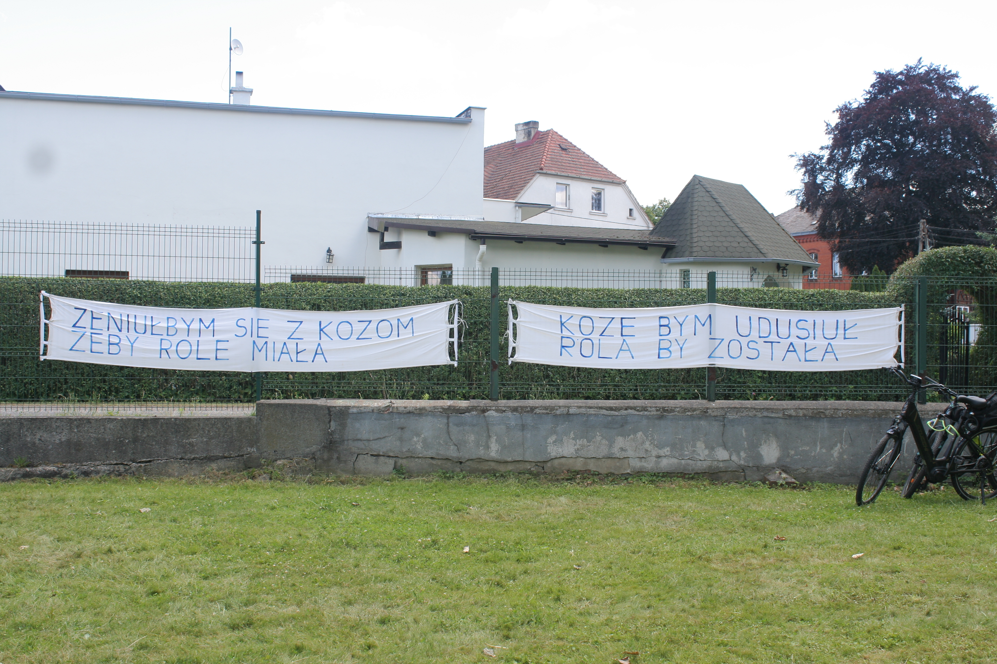Na zdjęciu widać dwa białe banery z niebieskimi napisami w języku polskim, rozwieszone na ogrodzeniu przed domem. Obok stoi rower.