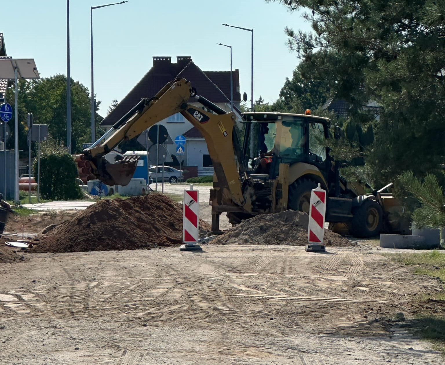 Na zdjęciu znajduje się koparka podczas prac ziemnych przy budowie ulicy Reymonta w Kole. Po prawej stronie widoczne są drzewa, a w tle zabudowania jednorodzinne.