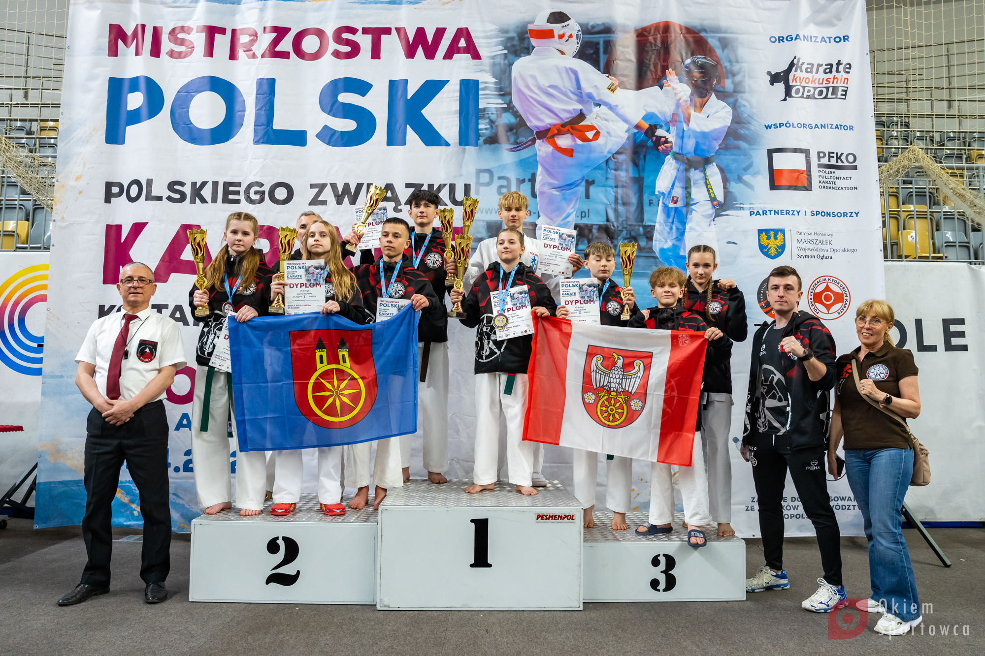 Kilkanaście zawodniczek i zawodników stoi na podium, trzymając puchary i dyplomy oraz flagi miasta Koło i Powiatu Kolskiego, obok nich stoją opiekunowie, a w tle widoczny jest duży baner z nazwą wydarzenia.