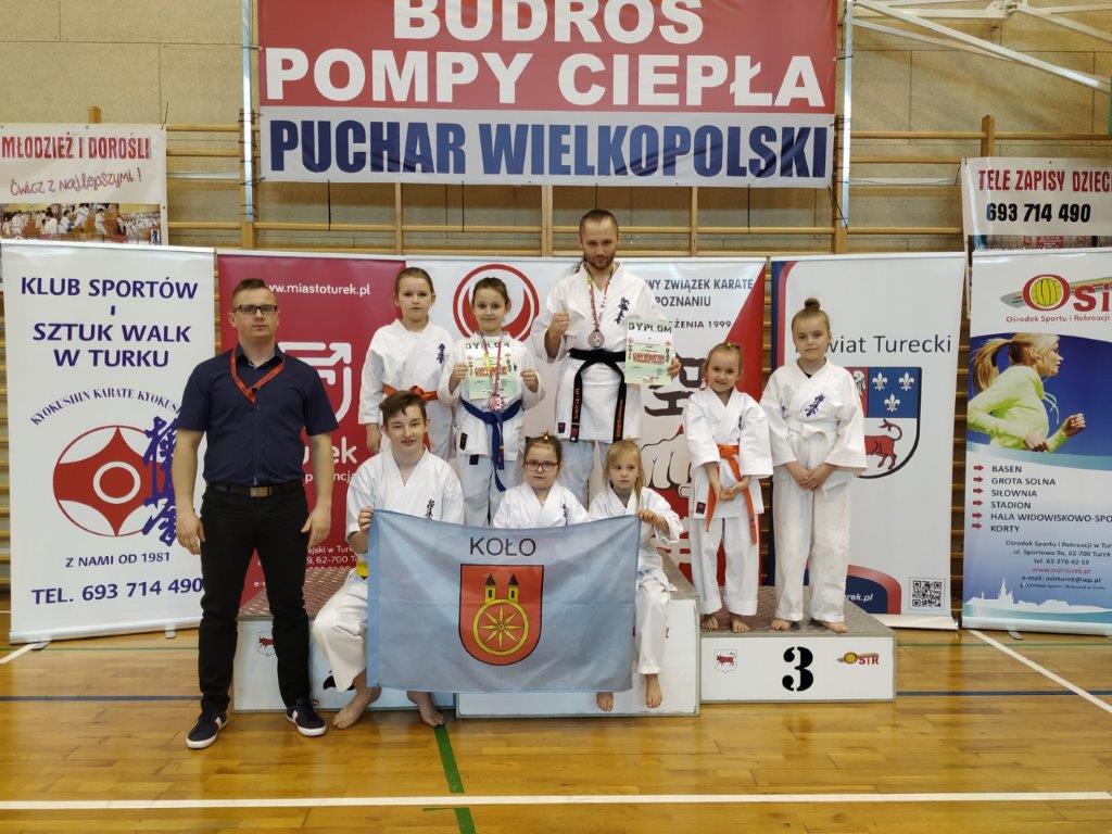 Kolscy karatecy stoją na podium, zawodnicy trzymają flagę miasta Koła.