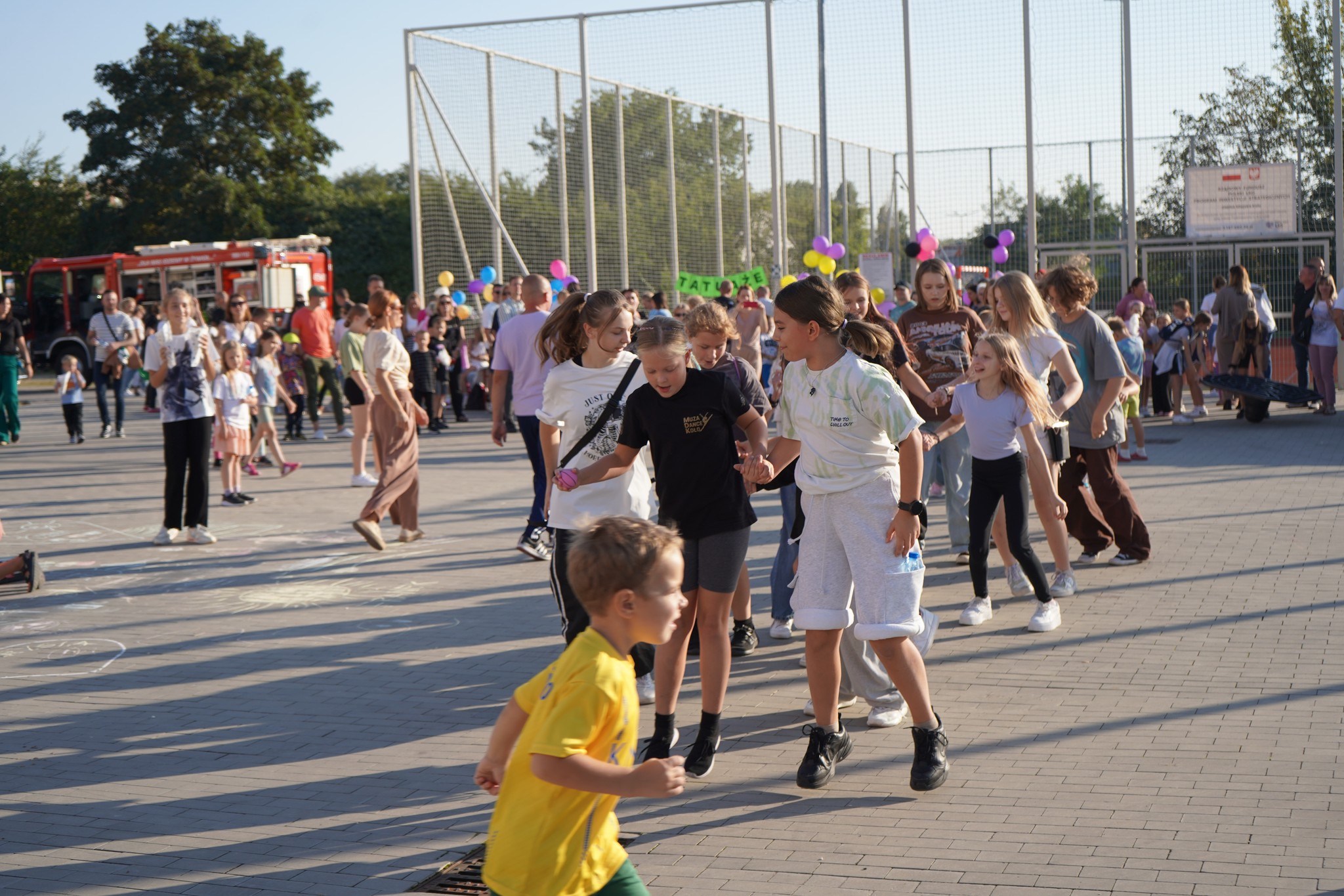W centrum zdjęcia widać grupę podskakujących dziewczynek i chłopców, stojących ramię w ramię, jedni za drugimi. Niektórzy mają na sobie krótkie spodenki i koszulki. W tle ogrodzone boisko, wóz strażacki, grupa spacerujących dzieci i dorosłych