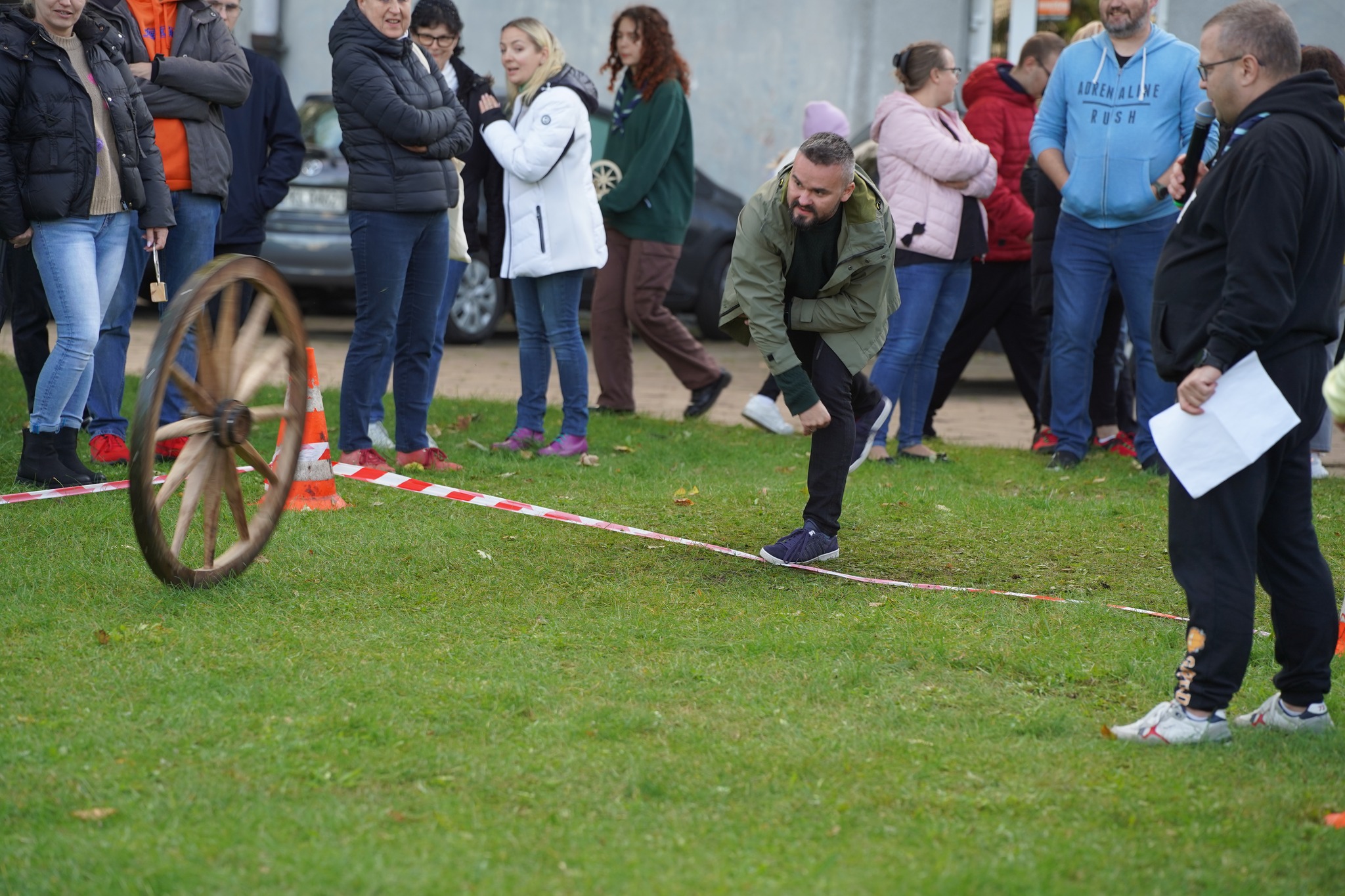 Mężczyzna toczy drewniane koło podczas zawodów plenerowych, obserwowany przez grupę uczestników i sędziego.