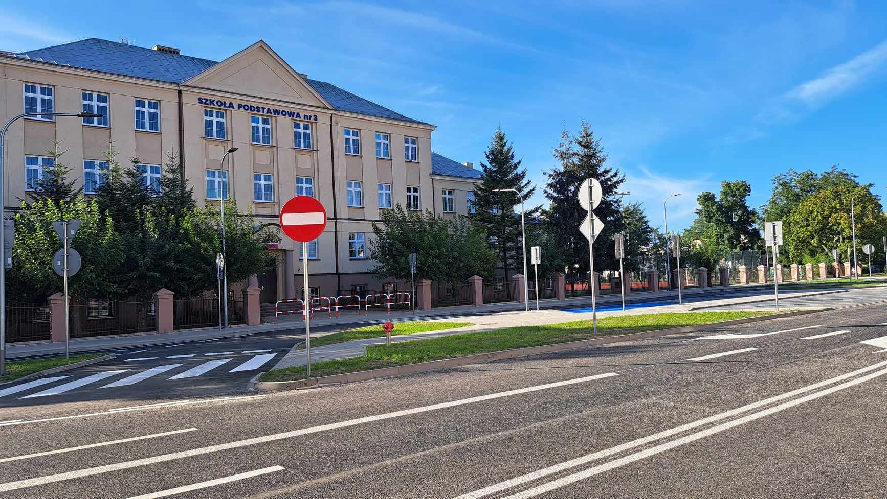 Na zdjęciu widać wyremontowaną ulicę w mieście Kole. W tle widać budynek szkoły podstawowej, przed nim ogrodzenie oraz drzewa.