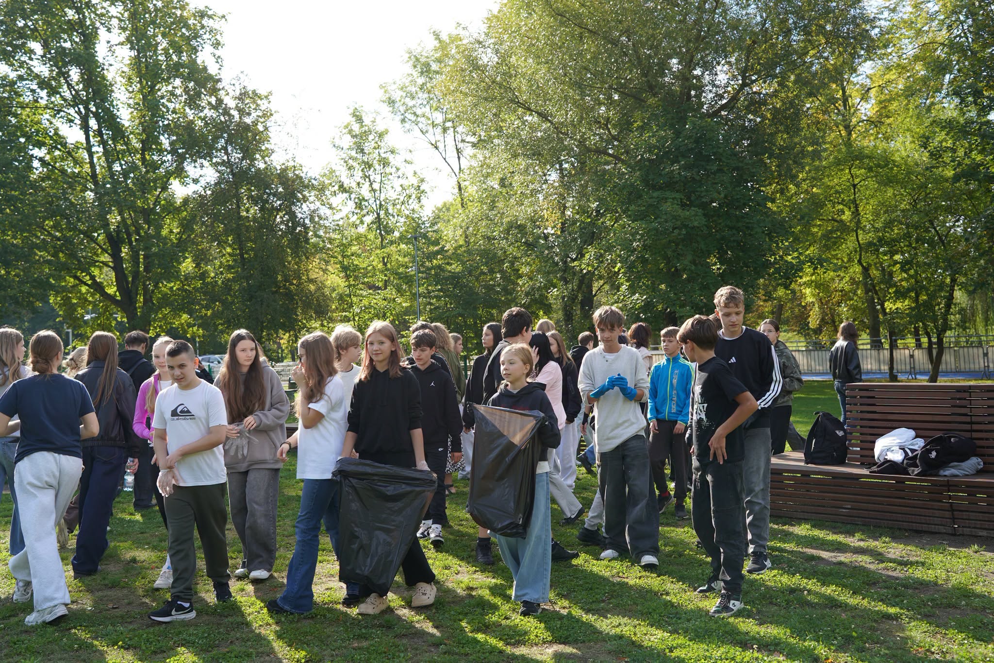 Grupa nastolatków sprząta park w słoneczny dzień. Dwie dziewczyny niosą duże czarne worki na śmieci, część uczestników ma założone rękawiczki ochronne. W tle zielone drzewa, drewniana ławka z plecakami oraz pozostali uczestnicy akcji sprzątania.