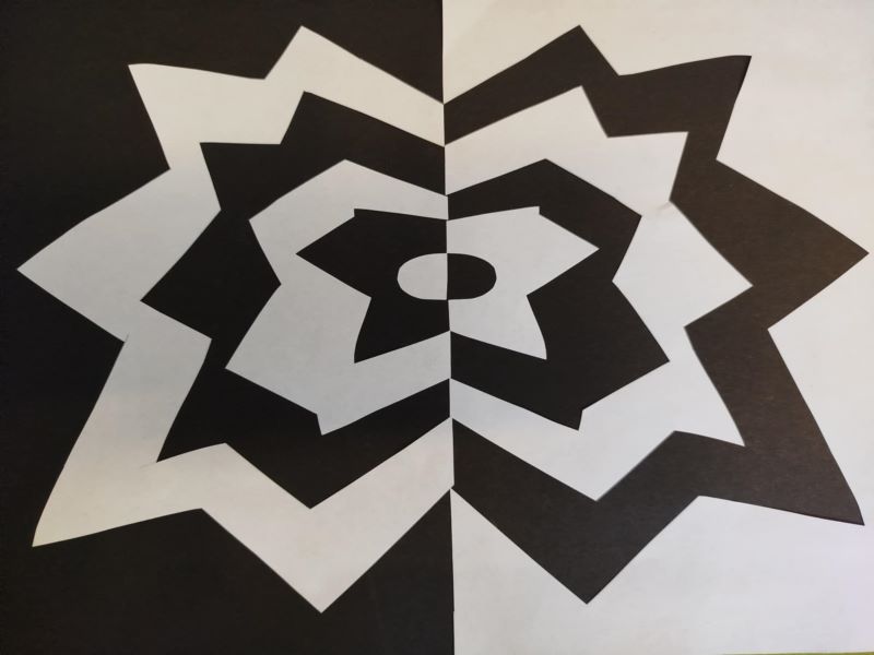 Grafika przedstawia symetryczny, czarno-biały wzór przypominający kwiat lub gwiazdę, z kontrastującymi kształtami ułożonymi naprzemiennie po obu stronach osi symetrii.