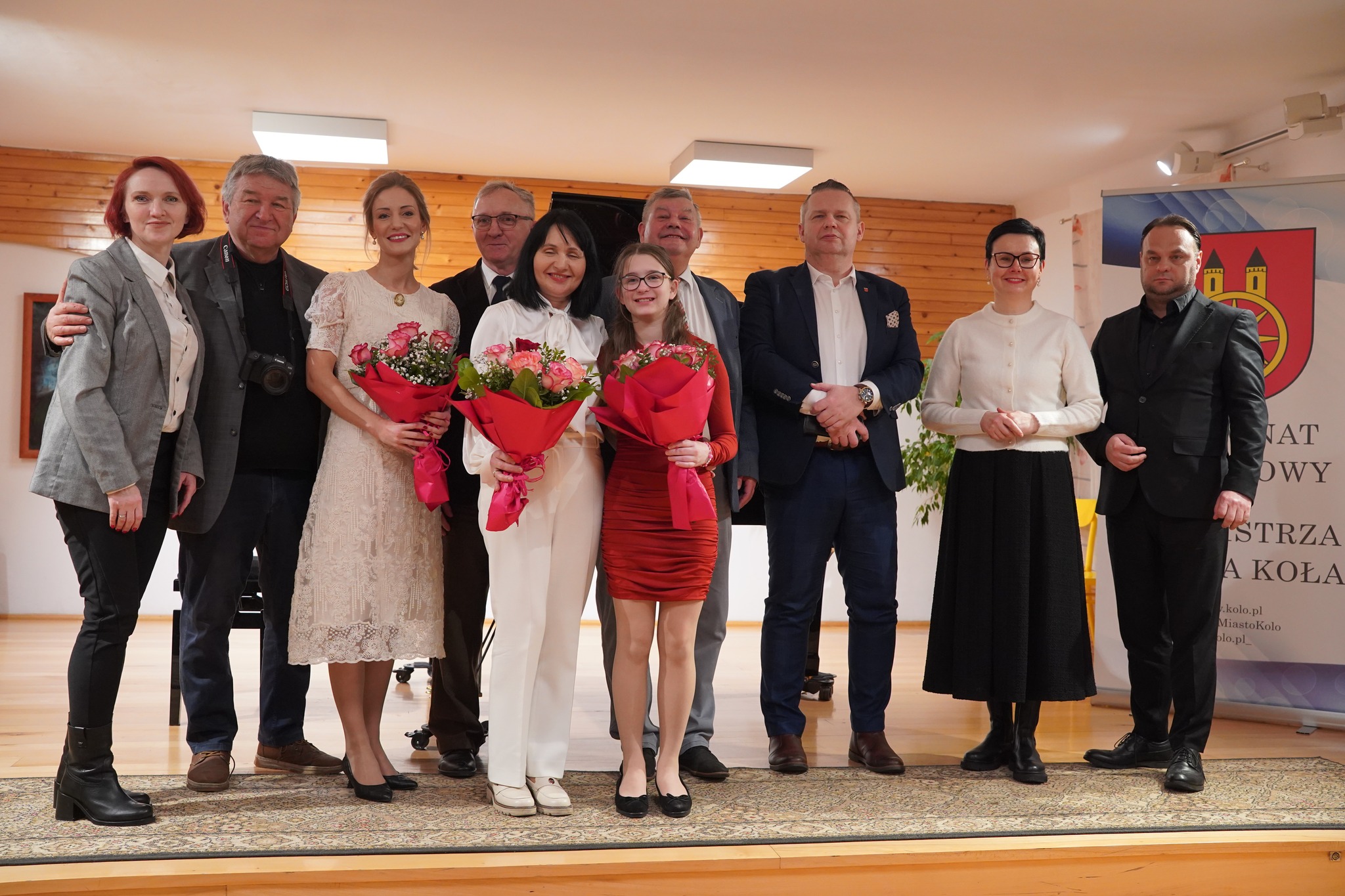 Na scenie w szkole muzycznej, na tle czarnego fortepianu, stoi grupa elegancko ubranych kobiet i mężczyzn, a w centrum dwie kobiety i dziewczynka trzymające w dłoniach kwiaty, co podkreśla uroczysty i wyjątkowy charakter tego wydarzenia.