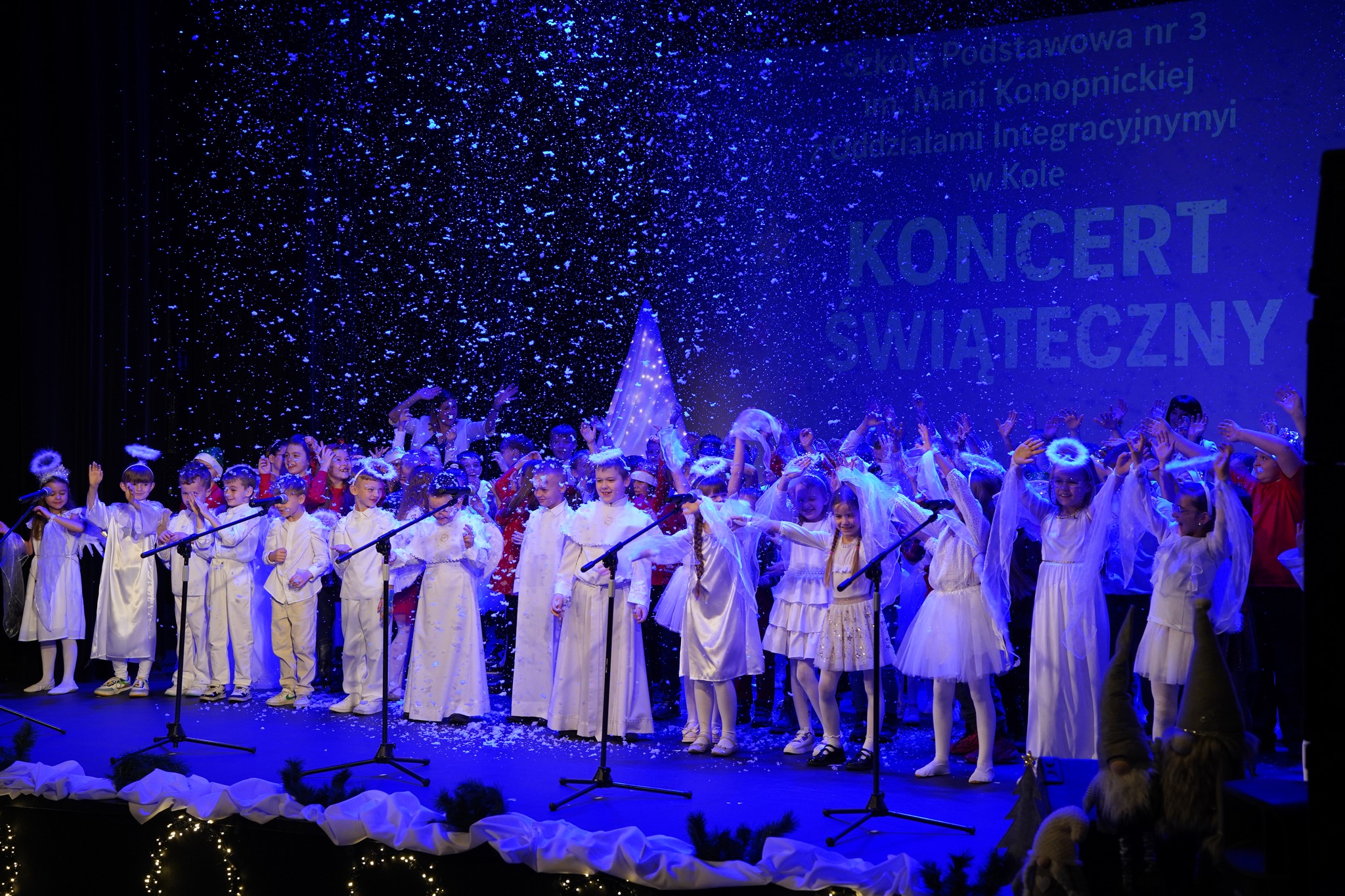 Na zdjęciu widać dzieci na scenie Miejskiego Domu Kultury, ubrane w stroje aniołów. Podczas występu unoszą dłonie w geście radości, a z góry spada konfetti. Przed nimi stoją statywy z mikrofonami, a w tle widoczny jest ekran kinowy z napisem Koncert 