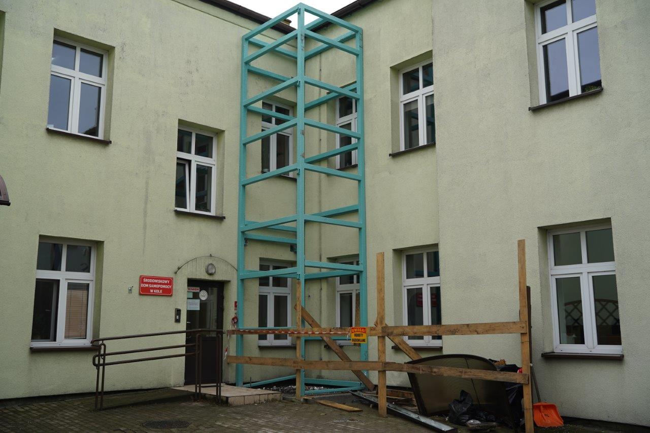 Na zdjęciu widać budynek gdzie trwają prace budowlane związane z montażem windy zewnętrznej. Na pierwszym planie widać zieloną stalową konstrukcję, zamontowaną od parteru po ostatnie piętro budynku.