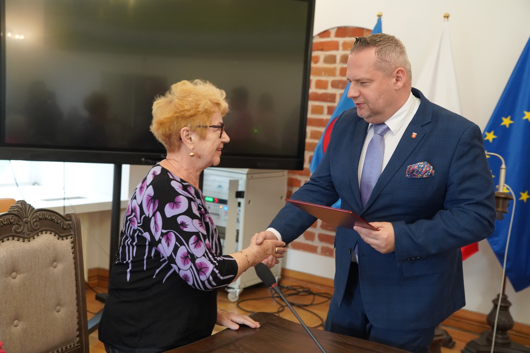 Na zdjęciu widać burmistrza Koła w garniturze, który przekazuje dokument dla seniorki, poświadczający odbiór opaski bezpieczeństwa. Spotkanie odbywa się w sali sesyjnej ratusza. W tle widać ekran oraz flagi miasta Koła, Polski oraz Unii Europejskiej.
