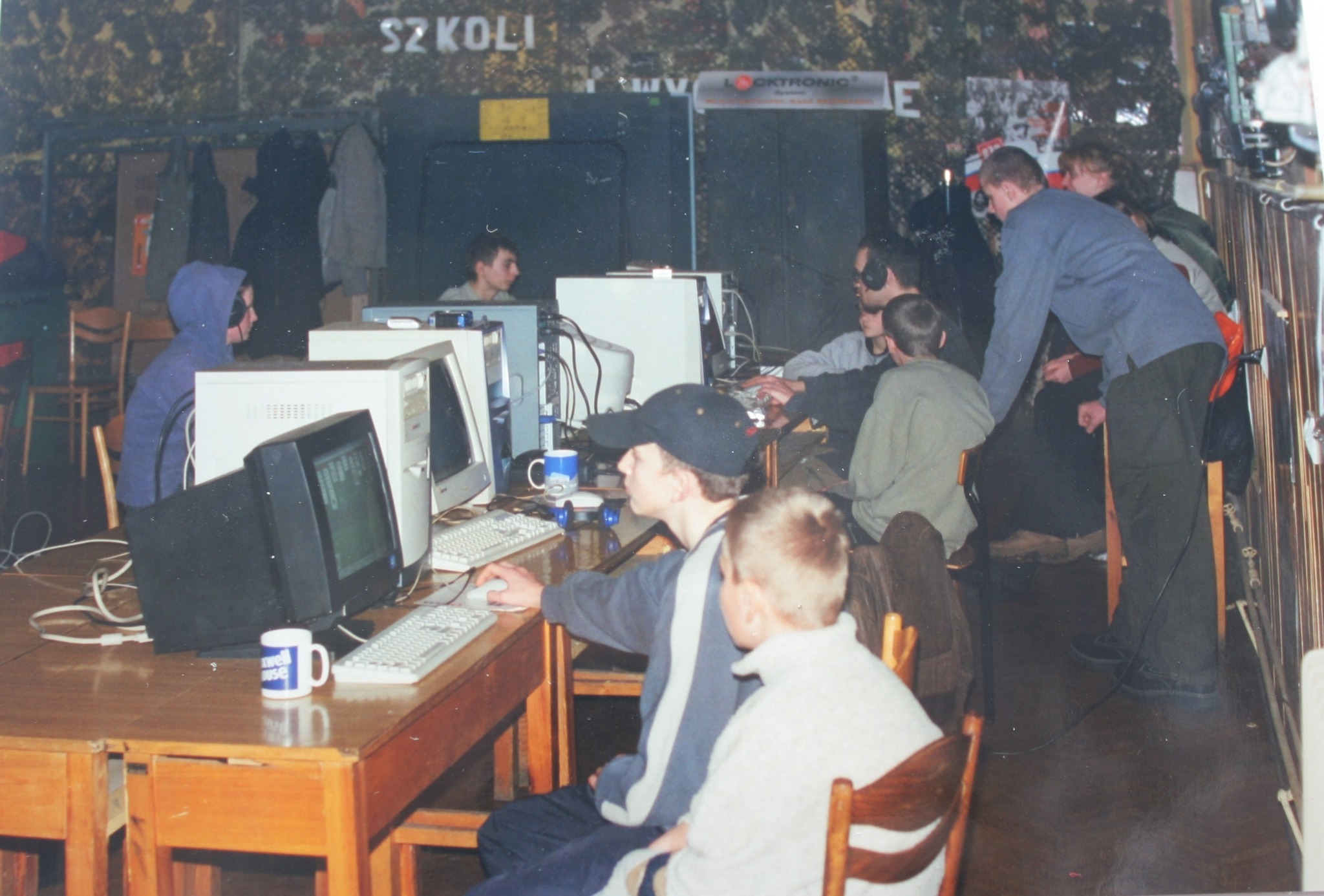 Zdjęcie przedstawia wnętrze pomieszczenia, w którym na stołach ustawione są monitory i stacje komputerowe, a przy nich siedzi i stoi młodzież. W tle widoczne są szafki i meble, a na ścianie zawieszony jest zielony element przypominający wojskowe wypo