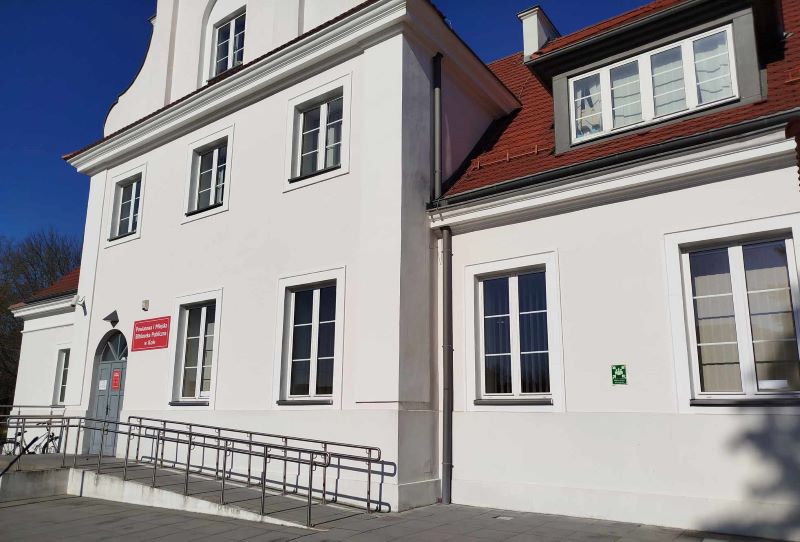 Biały, dwupiętrowy budynek użyteczności publicznej z czerwonym dachem i podjazdem dla osób z niepełnosprawnościami. Przy wejściu widoczna czerwona tablica z nazwą instytucji. Słoneczny dzień, błękitne niebo.