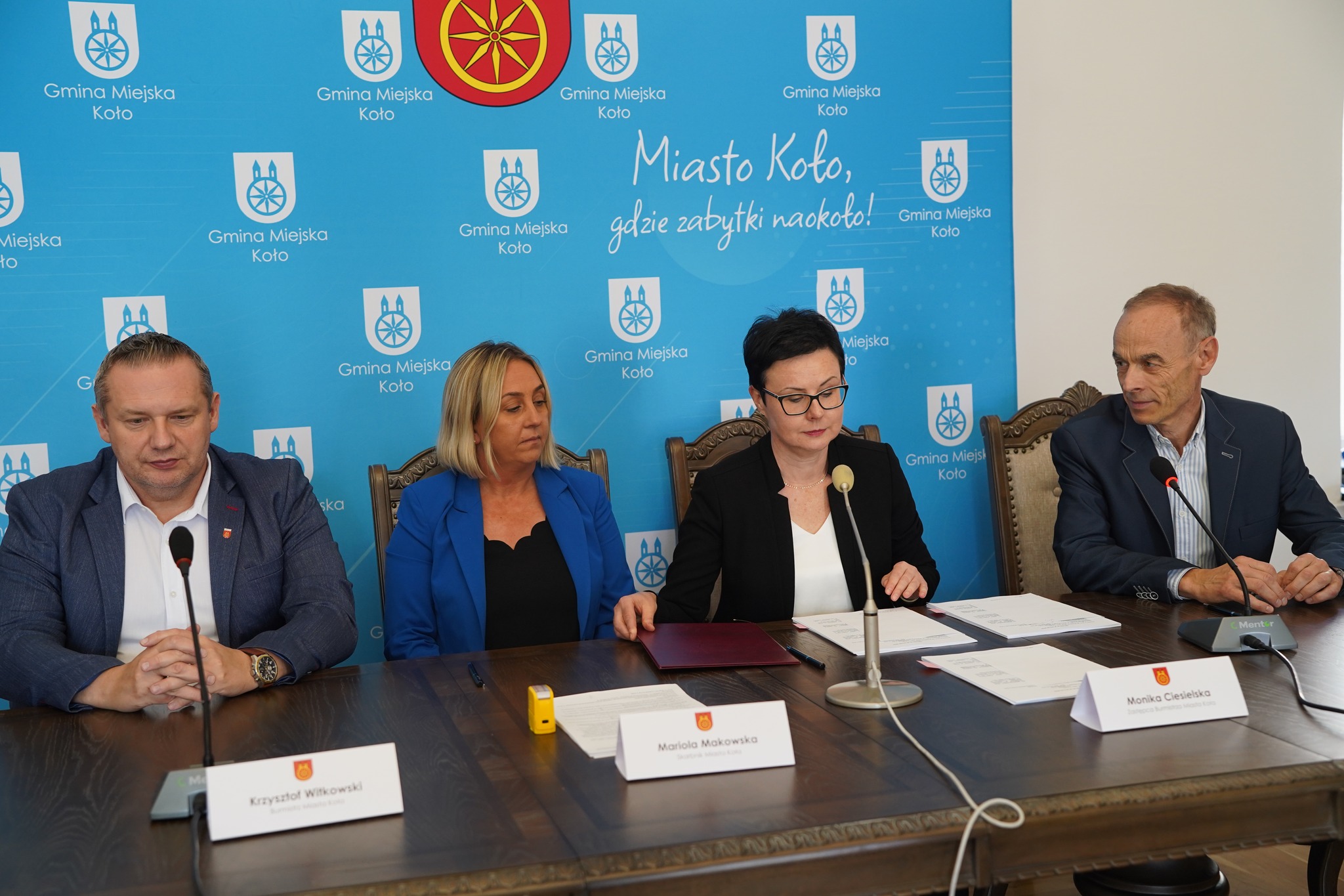 Na zdjęciu widać dwie kobiety oraz dwóch mężczyzn siedzących przy stole podczas oficjalnego podpisywania umowy, na tle ścianki promującej Gminę Miejską Koło z hasłem Miasto Koło gdzie zabytki naokoło.