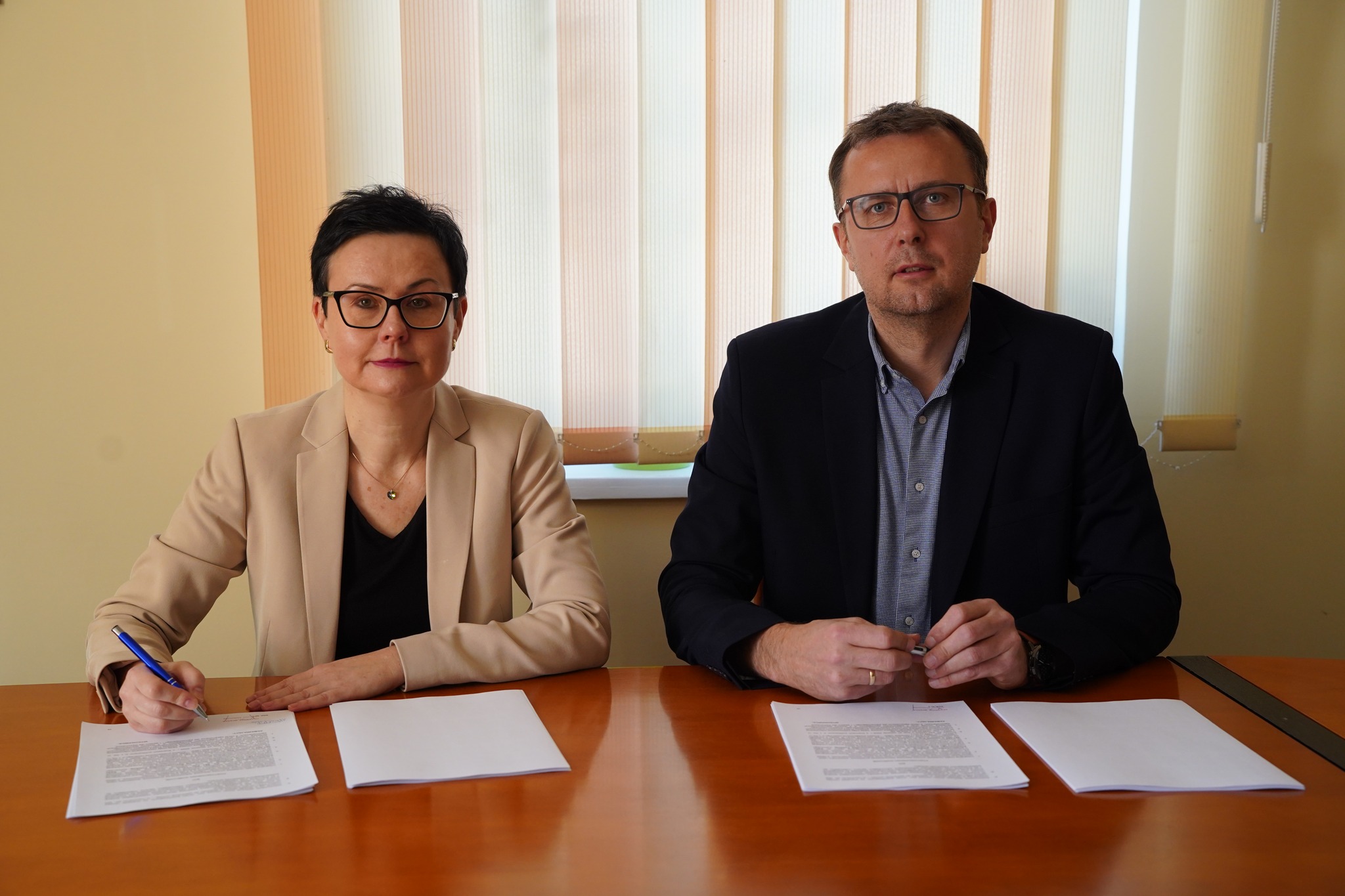 Na zdjęciu widać dwie osoby siedzące przy stole. Jedna z nich (po lewej, kobieta) podpisuje dokument, druga (po prawej, mężczyzna), trzyma w dłoniach, opartych na stole długopis. W tle widać okno z zasłoniętymi roletami.