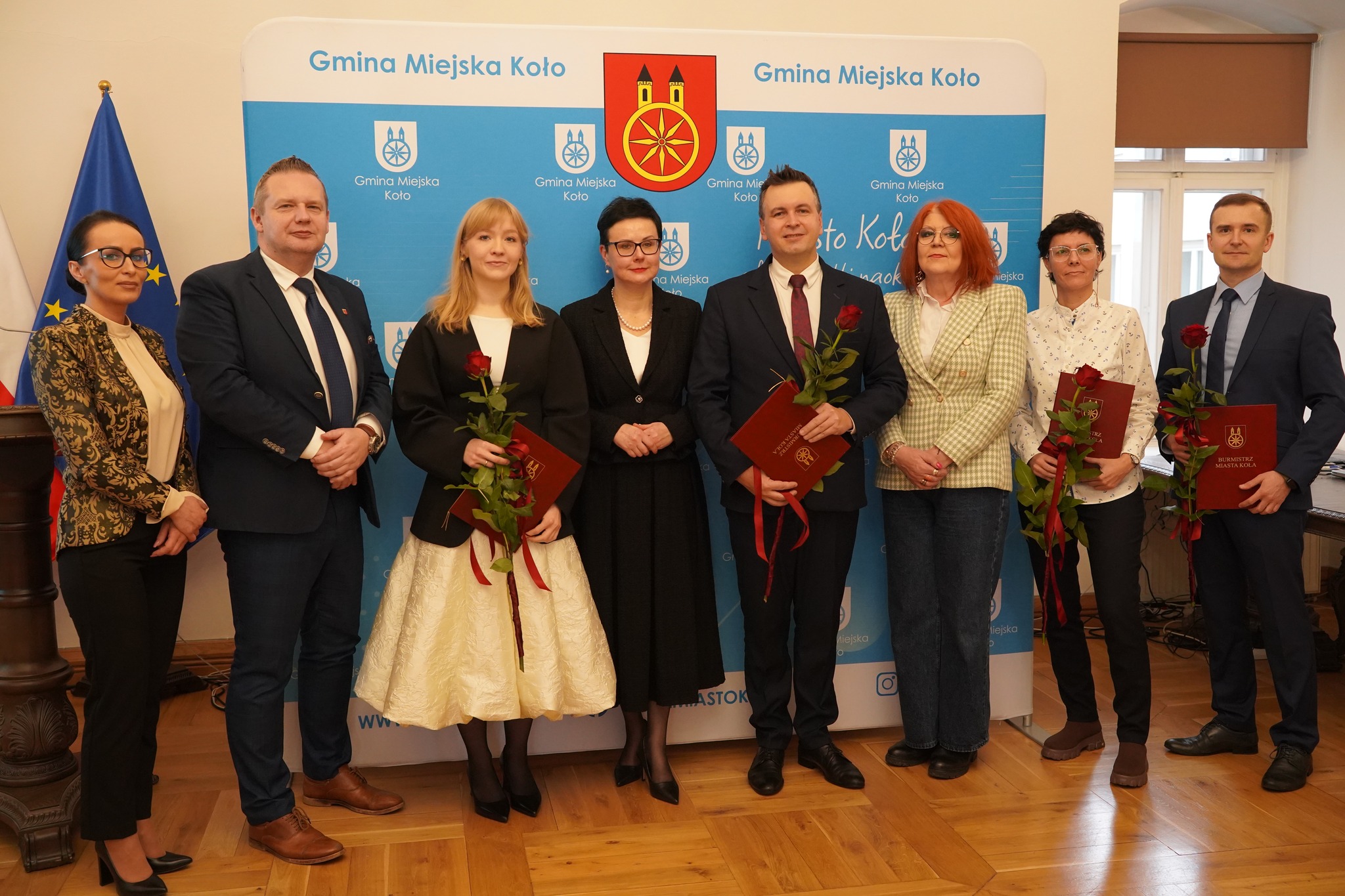 Na zdjęciu widać grupę ośmiu osób stojących w rzędzie podczas oficjalnego wydarzenia. Kilkoro z nich trzyma w rękach czerwone teczki i czerwone róże, co sugeruje wręczenie nagród lub wyróżnień. Wszyscy są elegancko ubrani i pozują do zdjęcia.