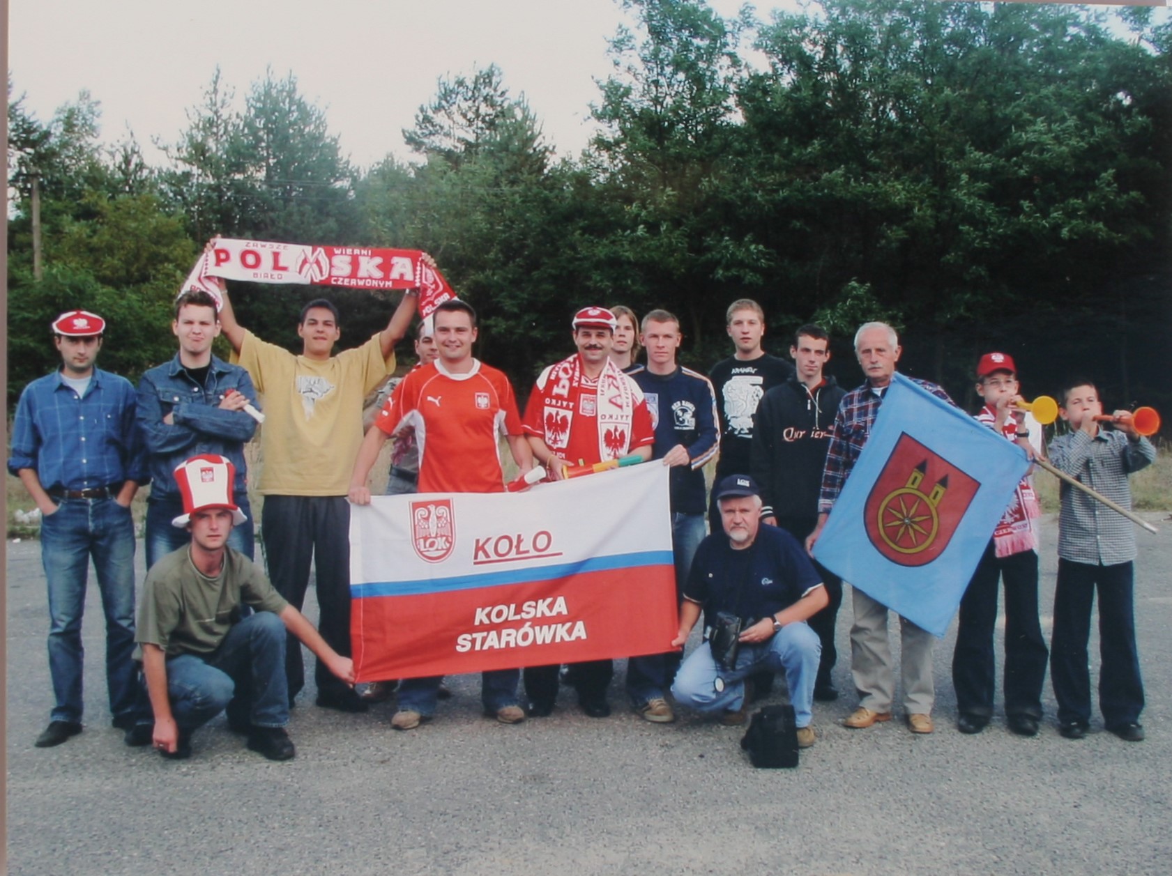Zdjęcie przedstawia grupę kibiców z Koła, wspierających polską drużynę. Ubrani są w kolorowe stroje, trzymają flagi i szaliki z napisami Polska oraz Koło Kolska Starówka. Inne niosą akcesoria kibicowskie: czapki i szaliki, a dzieci dmuchają w trąbki.