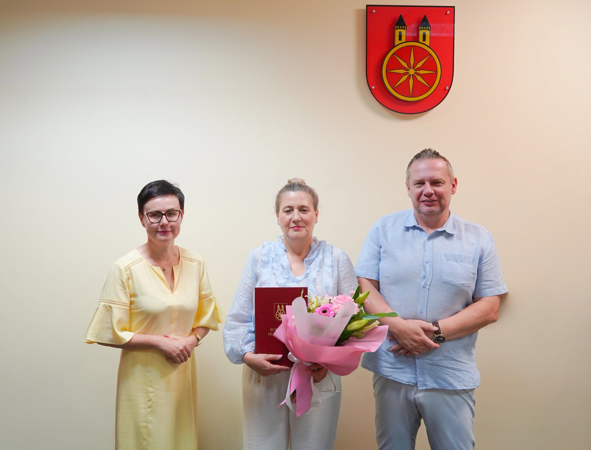 Troje osób stoi przy jasnej ścianie pod czerwonym herbem z żółtym kołem i dwiema wieżami. Kobieta w środku trzyma bordową teczkę z herbem i bukiet różowych kwiatów. Po lewej kobieta w żółtej sukience, po prawej mężczyzna w błękitnej koszuli.