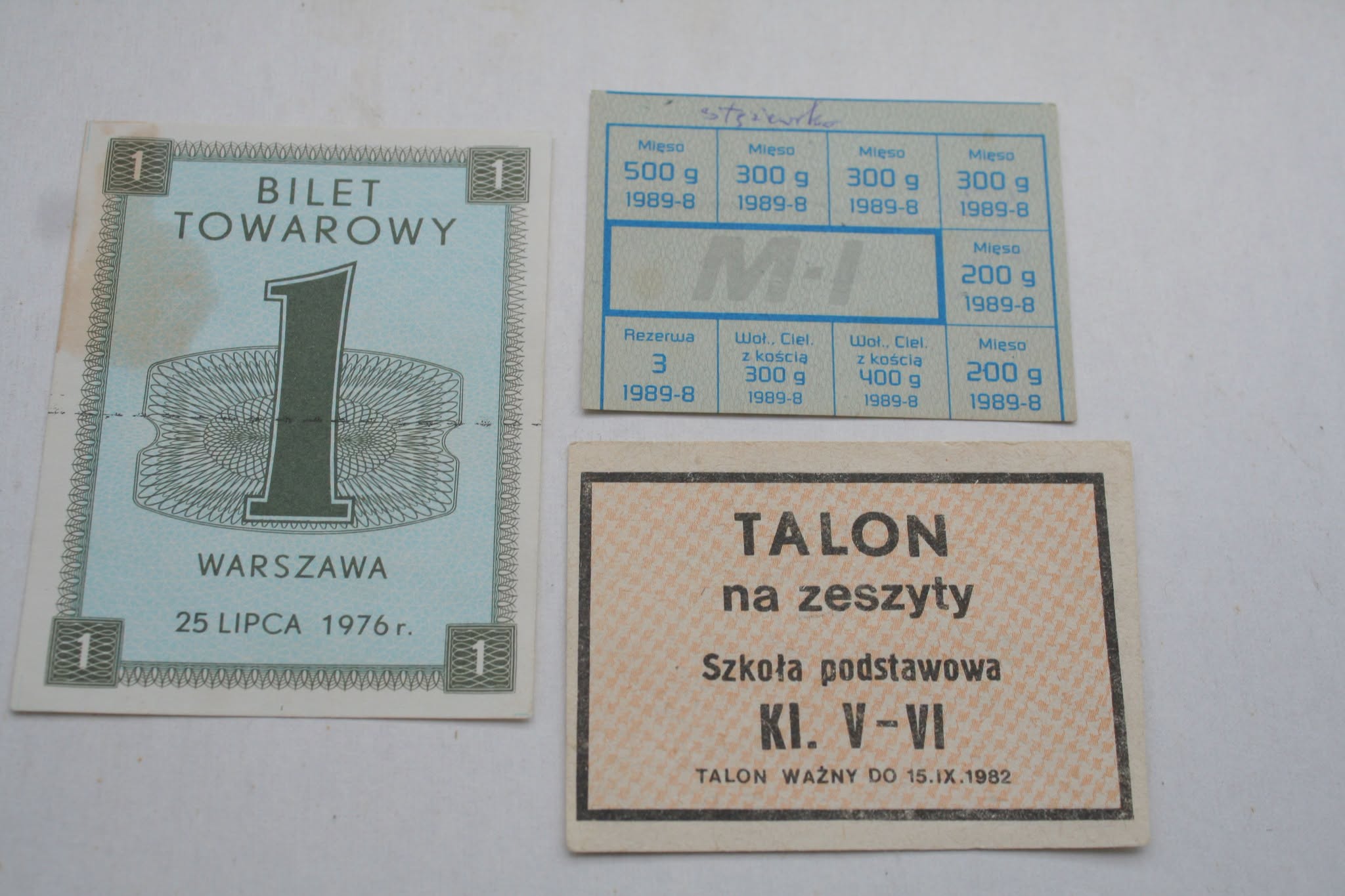 Trzy polskie kartki reglamentacyjne z czasów PRL na jasnym tle: niebieski Bilet Towarowy 1, Warszawa, 25 lipca 1976, kartka na mięso z 1989 z gramaturami oraz pomarańczowy Talon na zeszyty dla szkoły podstawowej kl. V-VI, ważny do 1982.