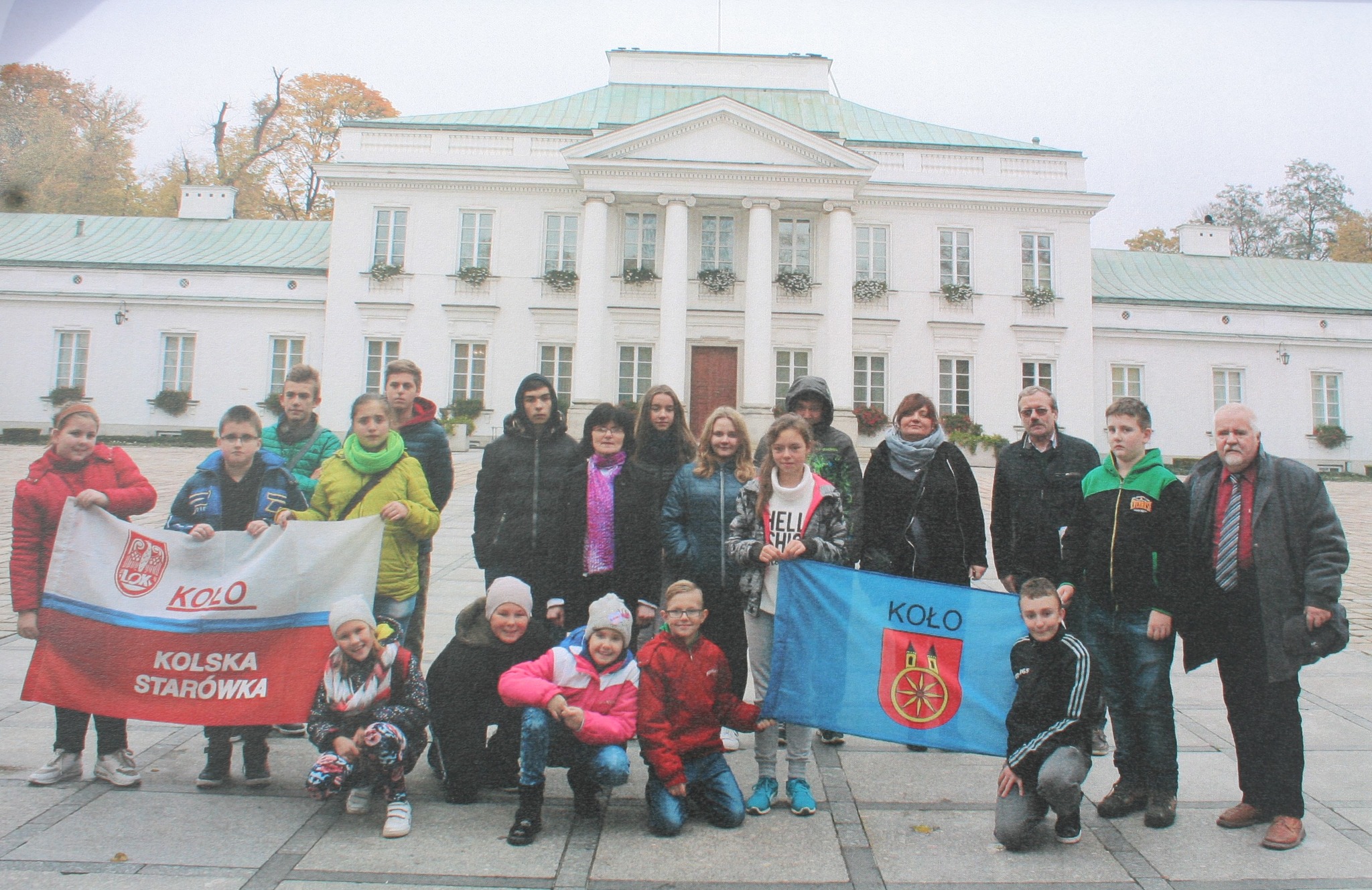 Na zdjęciu widać grupę osób, głównie dzieci i młodzież, stojących przed białym pałacem, trzymających flagi z napisem „Koło” i „Kolska Starówka”