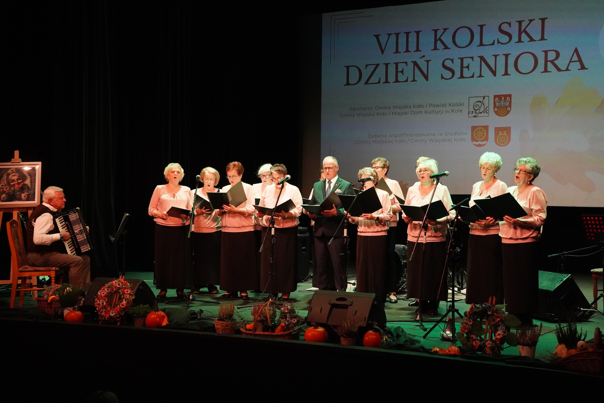 Na scenie występuje chór seniorów z akompaniamentem akordeonu podczas VIII Kolskiego Dnia Seniora w MDK w Kole.