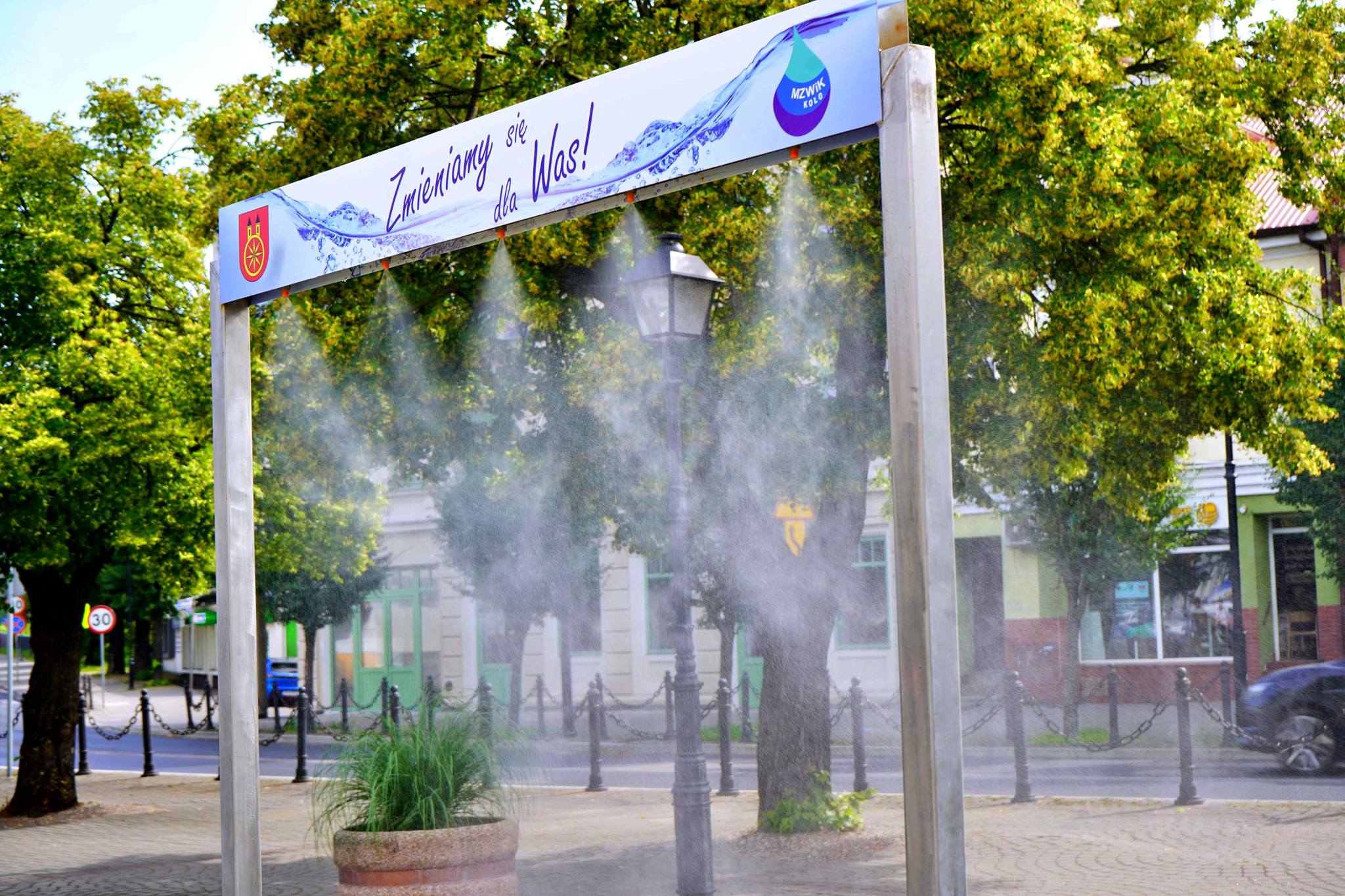Metalowa rama z kurtyna wodną mgiełkową ustawiona na rynku miejskim. Na górze baner z napisem Zmieniamy się dla Was oraz logo MZWIK Koło. W tle drzewa i zabytkowa zabudowa.