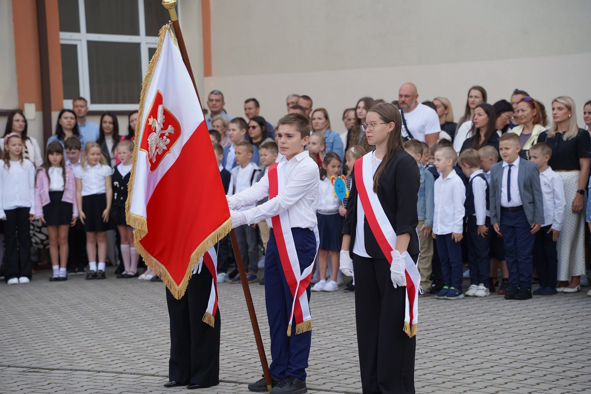 Uroczystość szkolna na dziedzińcu. Poczet sztandarowy — uczniowie w białych koszulach, biało-czerwonych szarfach i białych rękawiczkach — trzyma sztandar z godłem Polski. W tle stoją w szeregu młodsi uczniowie oraz zgromadzeni rodzice i nauczyciele.