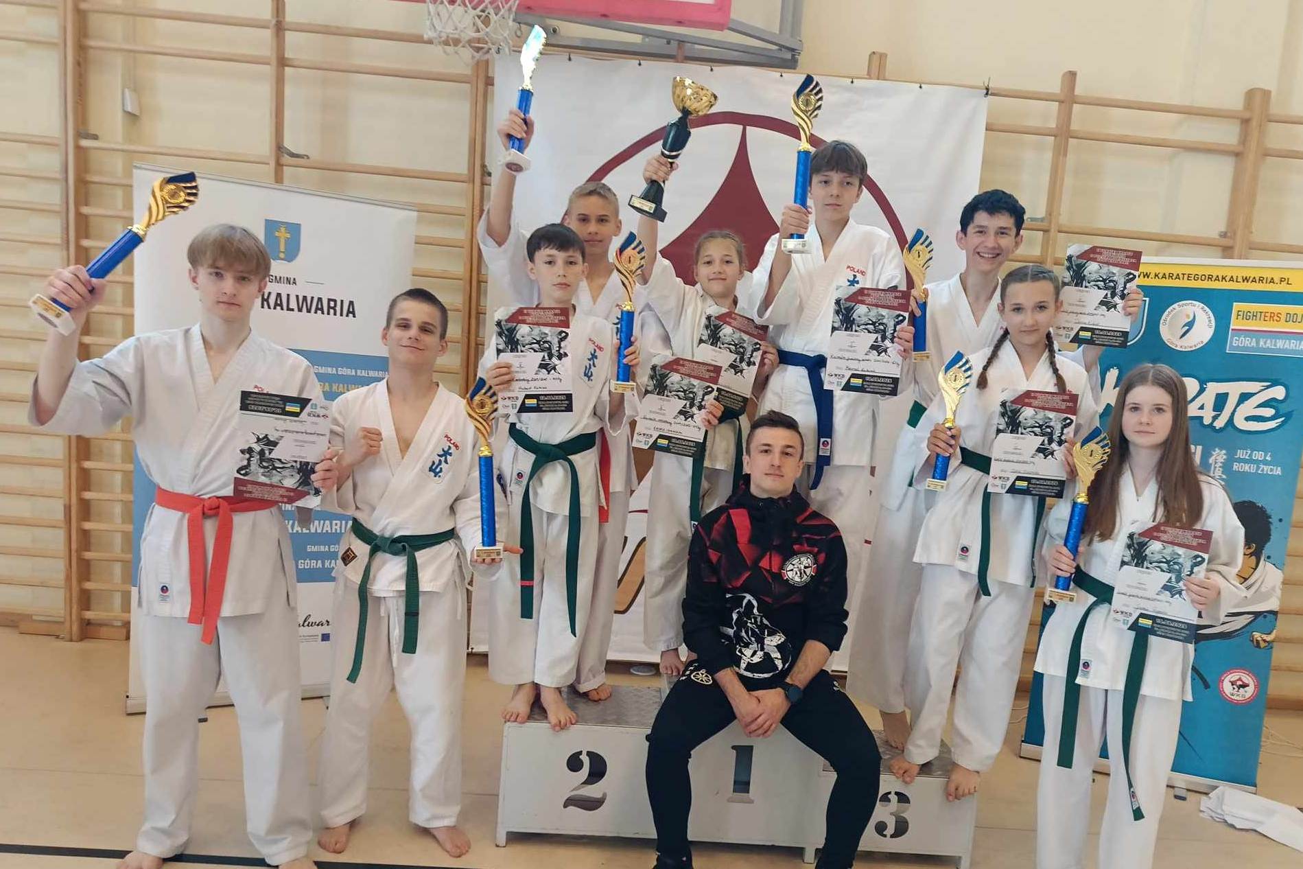 Grupa młodych karateków, dziewcząt i chłopców z dyplomami i medalami pozuje do zdjęcia w hali sportowej wraz z opiekunem. Za nimi widać drabinki do ćwiczeń, na nich wiszący baner oraz kosz do koszykówki.