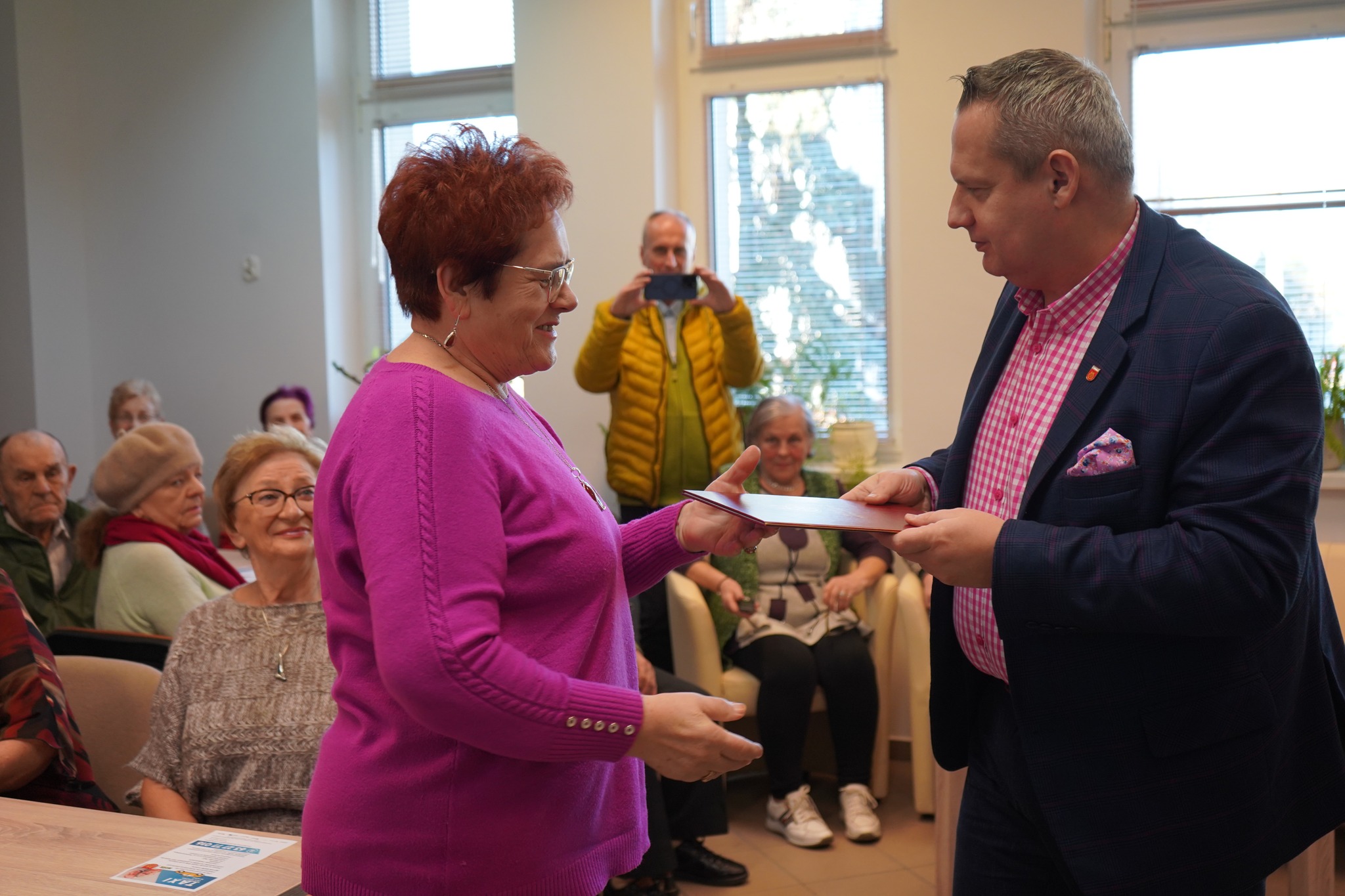 Na zdjęciu widać burmistrza Koła w marynarce i różowej koszuli, który przekazuje dokument dla seniorki. W tle znajdują się seniorzy siedzący na krzesłach. Jeden z uczestników robi zdjęcie, trzymając telefon.