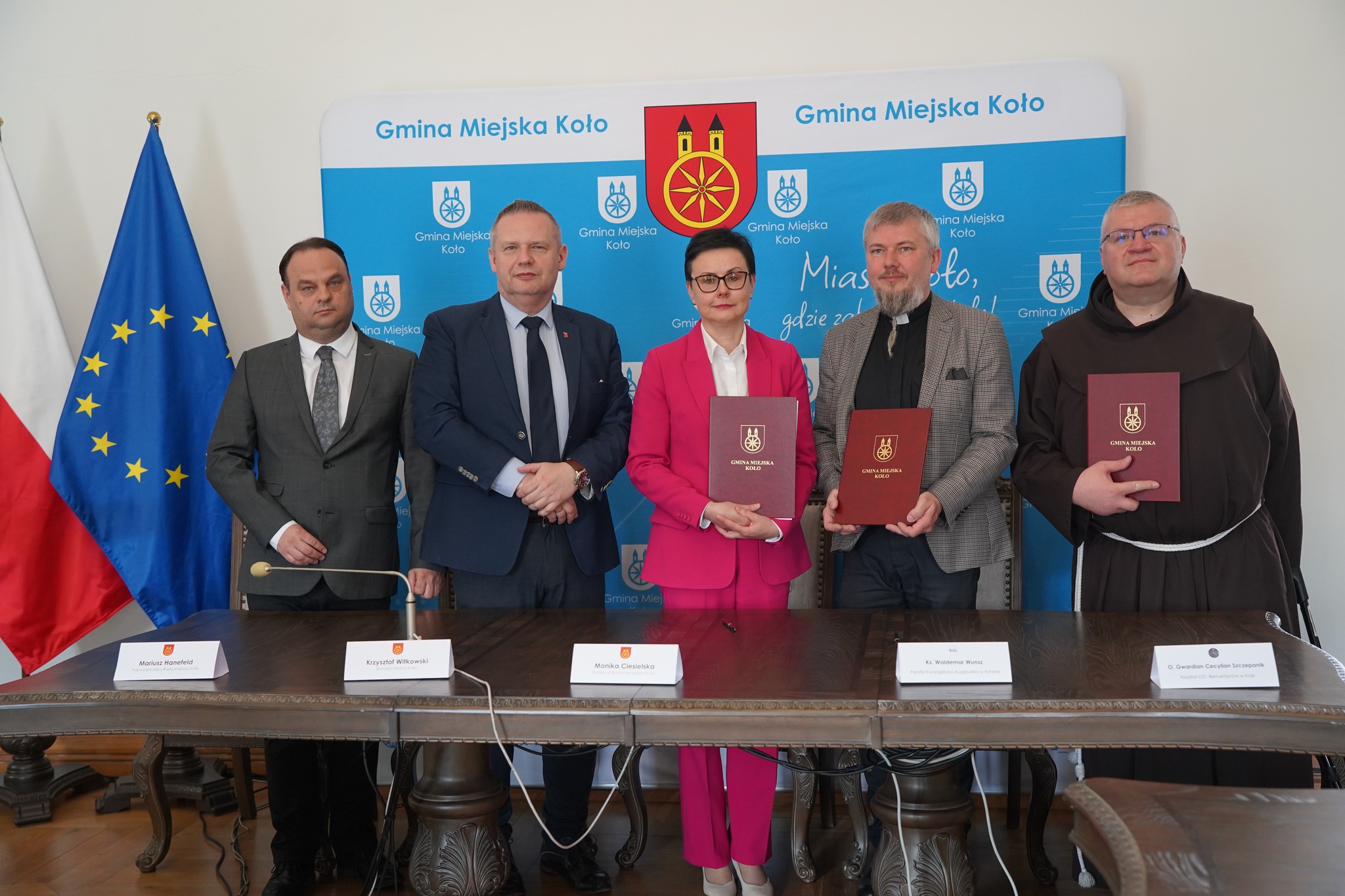 Na zdjęciu pięć osób – czterech mężczyzn i jedna kobieta – stoi na tle ścianki promocyjnej „Gmina Miejska Koło – Miasto Koło, gdzie zabytki naokoło”. Trzy osoby trzymają eleganckie teczki z herbem miasta Koła.