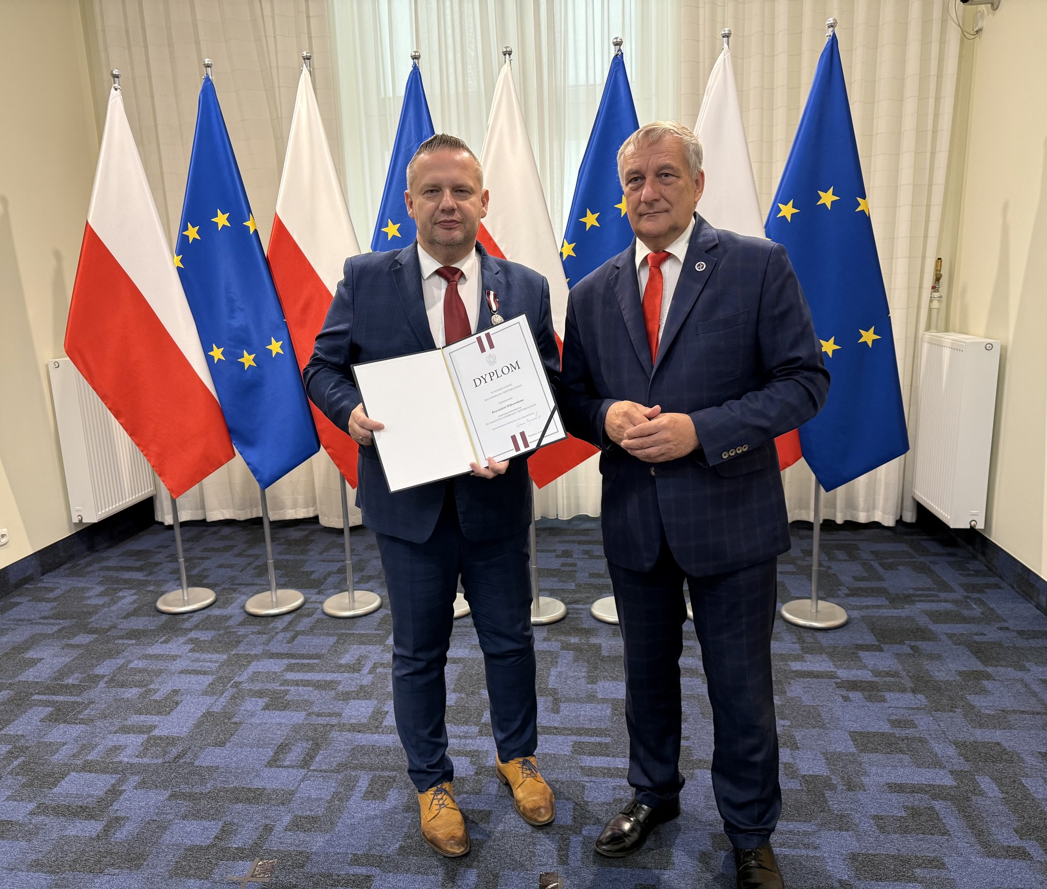 Na zdjęciu dwóch mężczyzn w garniturach pozuje z dyplomem na tle flag Polski i Unii Europejskiej – jeden z nich otrzymał wyróżnienie, burmistrz miasta Koła
