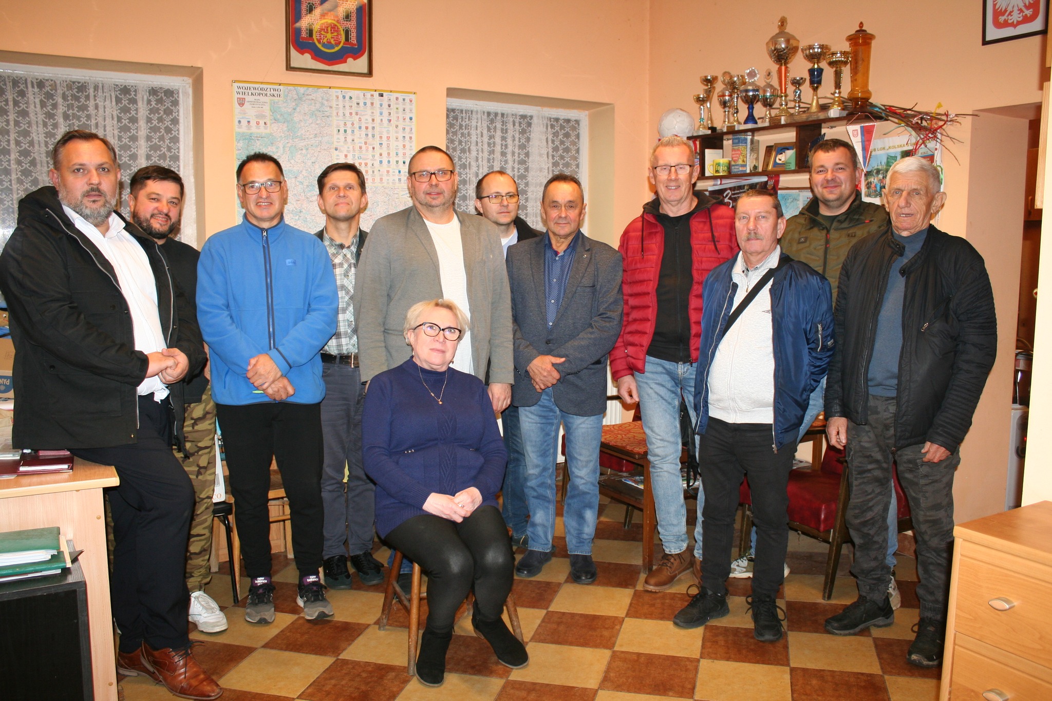 Na zdjęciu grupa osób pozuje wspólnie w pomieszczeniu, prawdopodobnie podczas spotkania lub zebrania. Jedna kobieta siedzi na pierwszym planie, pozostali stoją za nią.