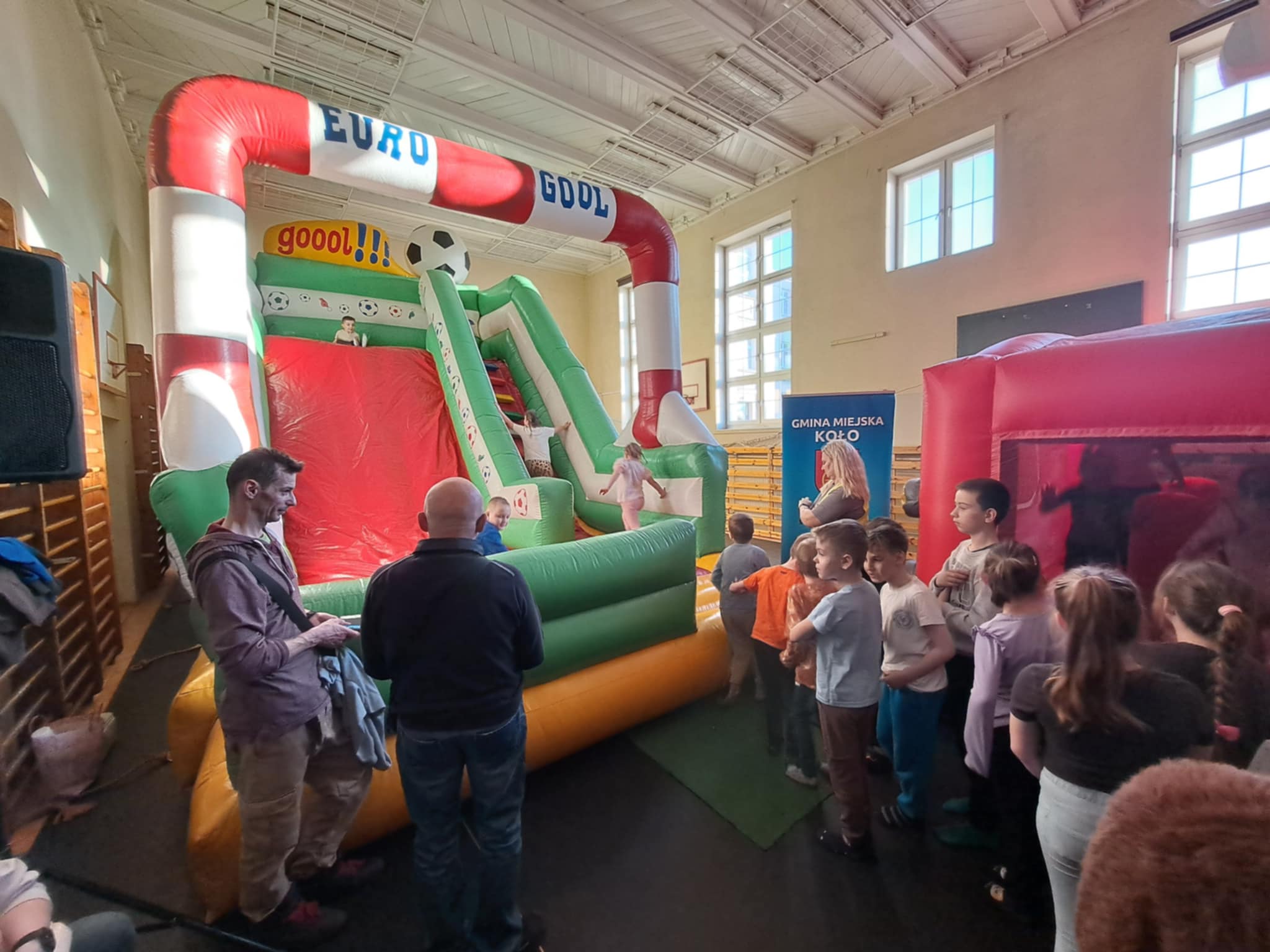 Zdjęcie przedstawia dzieci bawiące się na kolorowych dmuchanych atrakcjach w sali gimnastycznej. W tle widać opiekunów oraz baner z herbem miasta Koło i napisem Gmina Miejska Koło. Atmosfera jest radosna.
